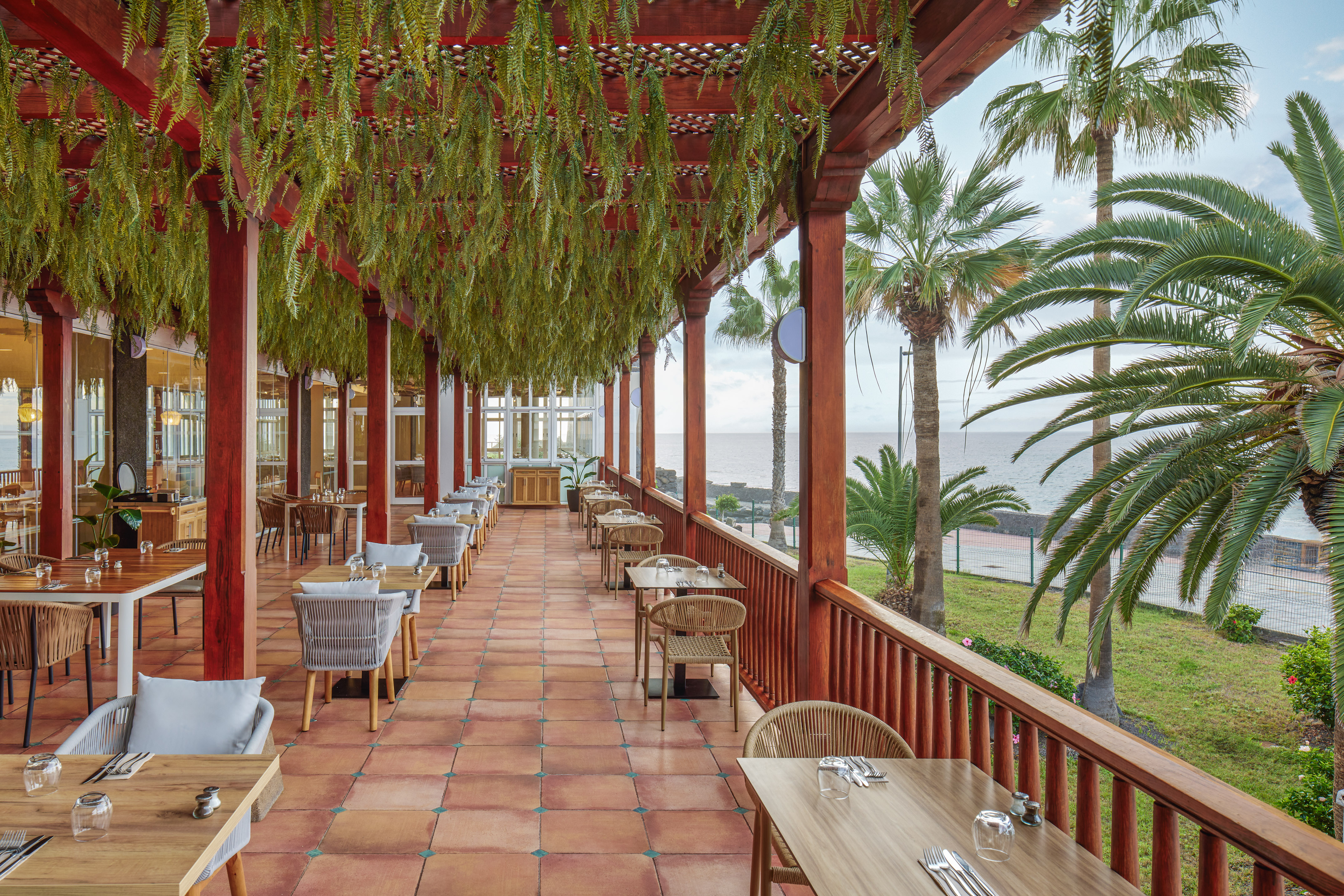 Outdoor restaurant terrace with ocean view, palm trees, and hanging ferns.