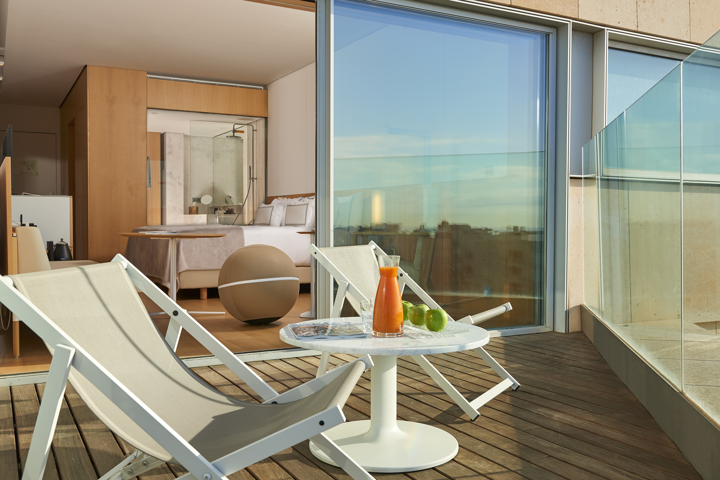 Modern hotel suite with balcony, deck chairs, fresh juice, and city views.
