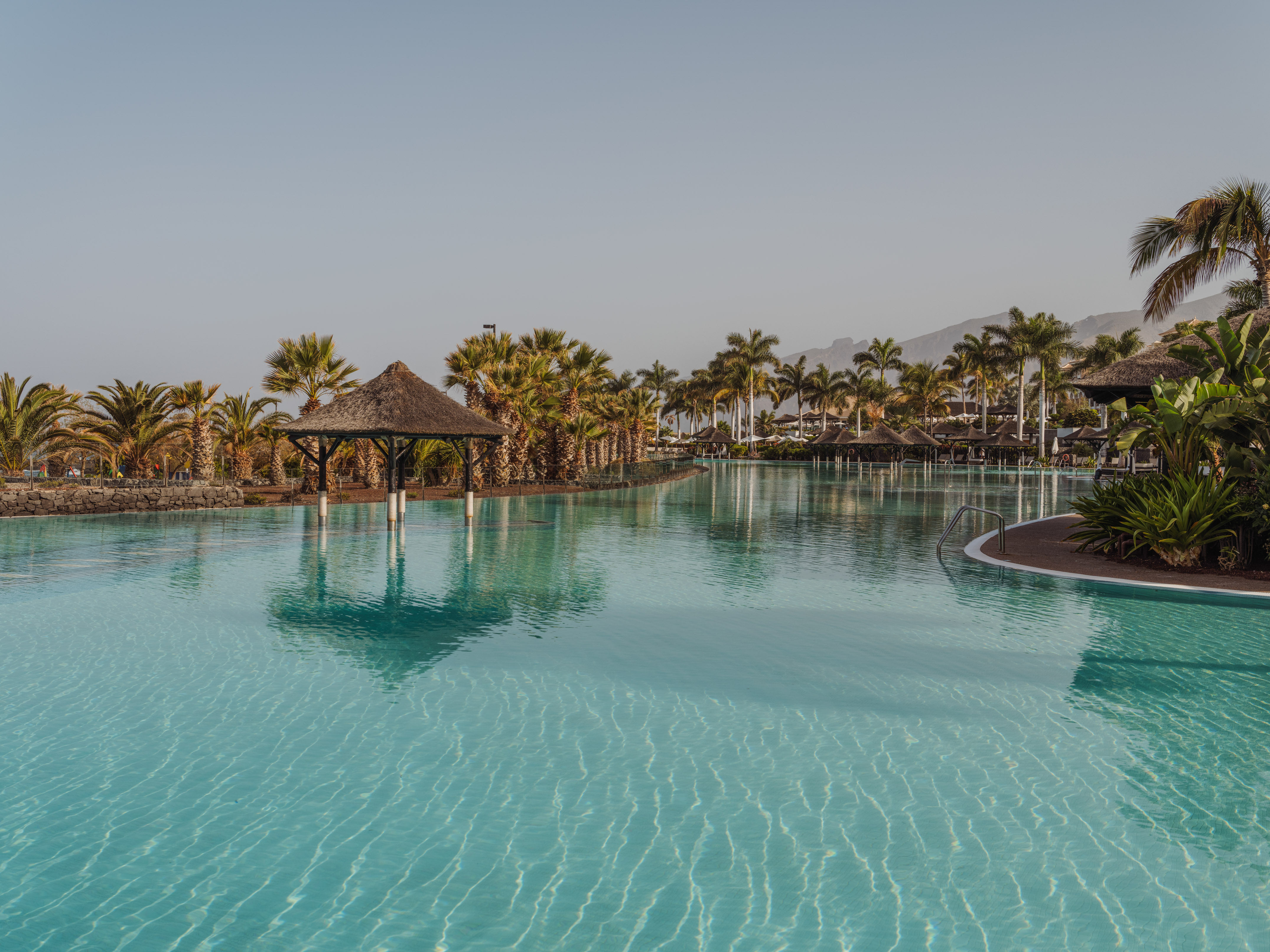 Blue water resort pool, palm trees, thatched gazebos, and distant mountains.