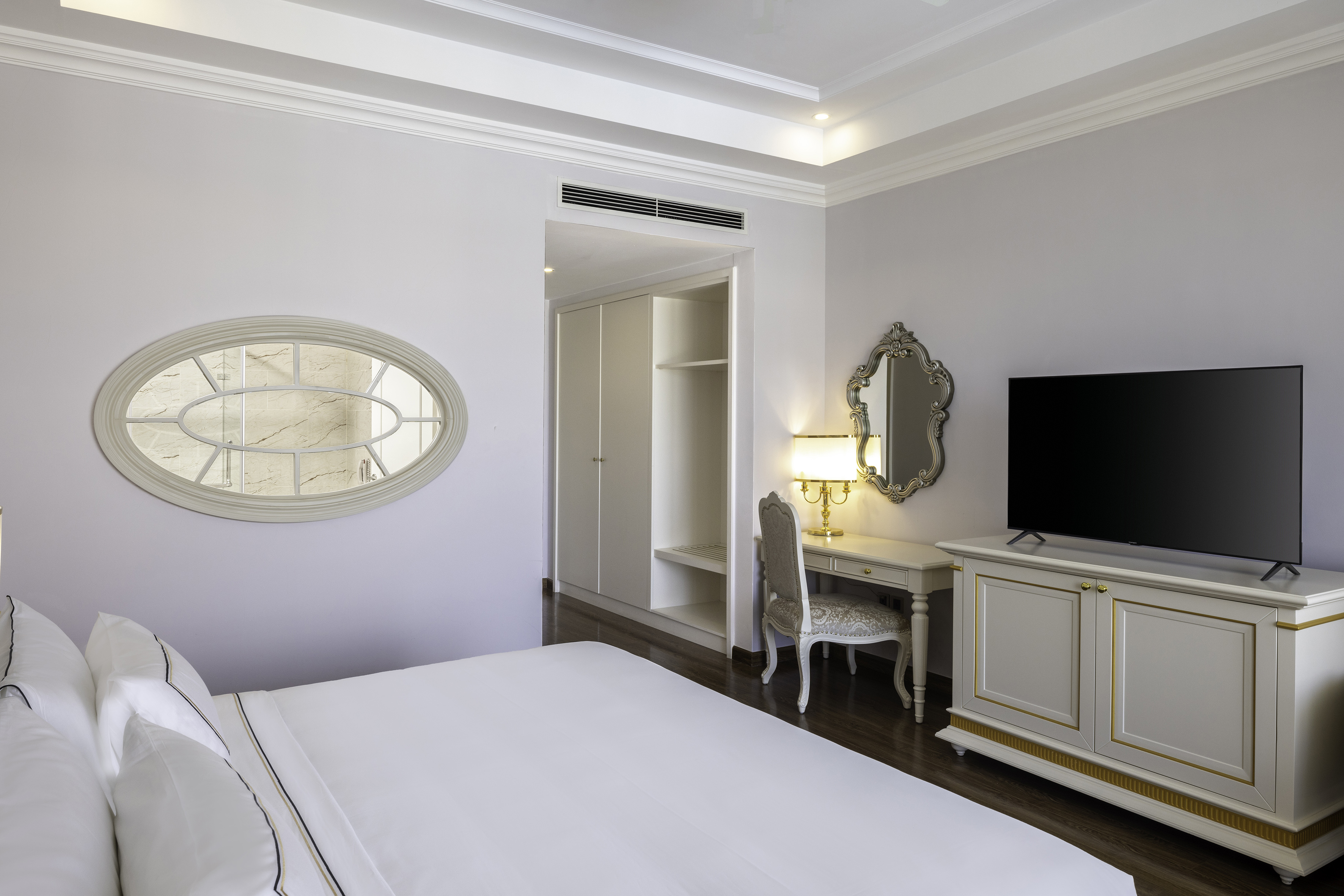 Elegant hotel bedroom with white bed, TV, dressing table, and decorative mirrors.