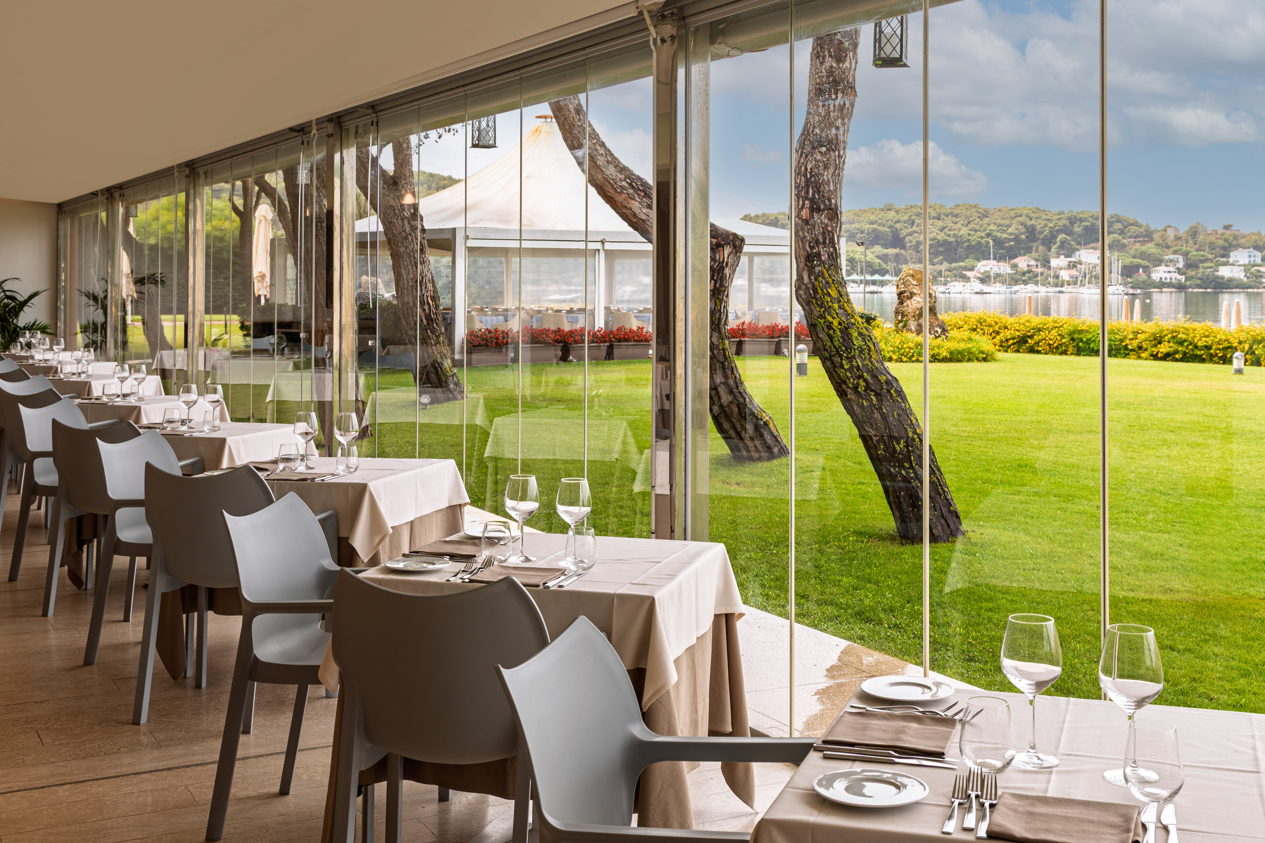 Restaurant dining room with tables, glass walls, and a scenic garden waterfront view.