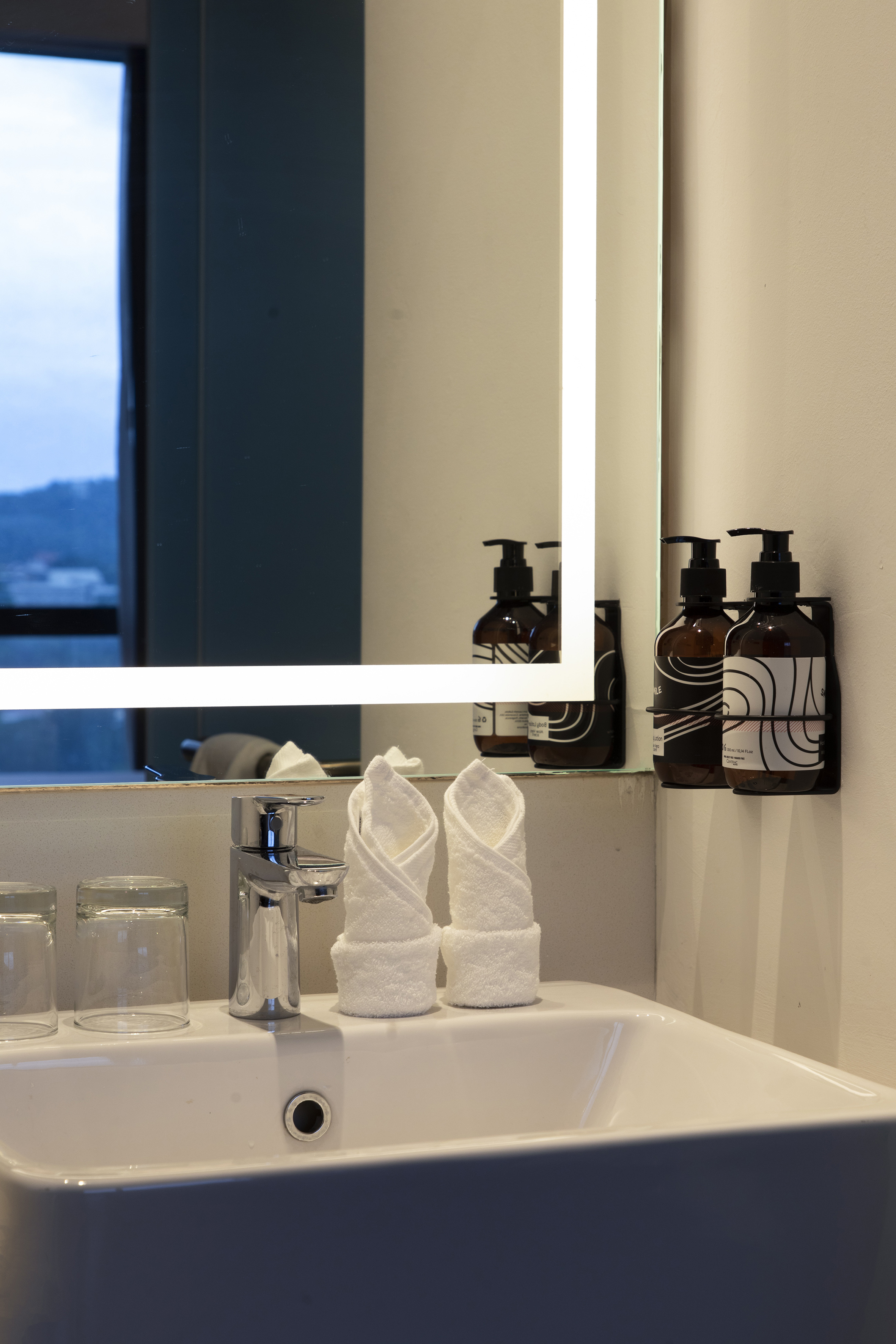 Modern bathroom sink, chrome faucet, rolled towels, toiletries, backlit mirror.
