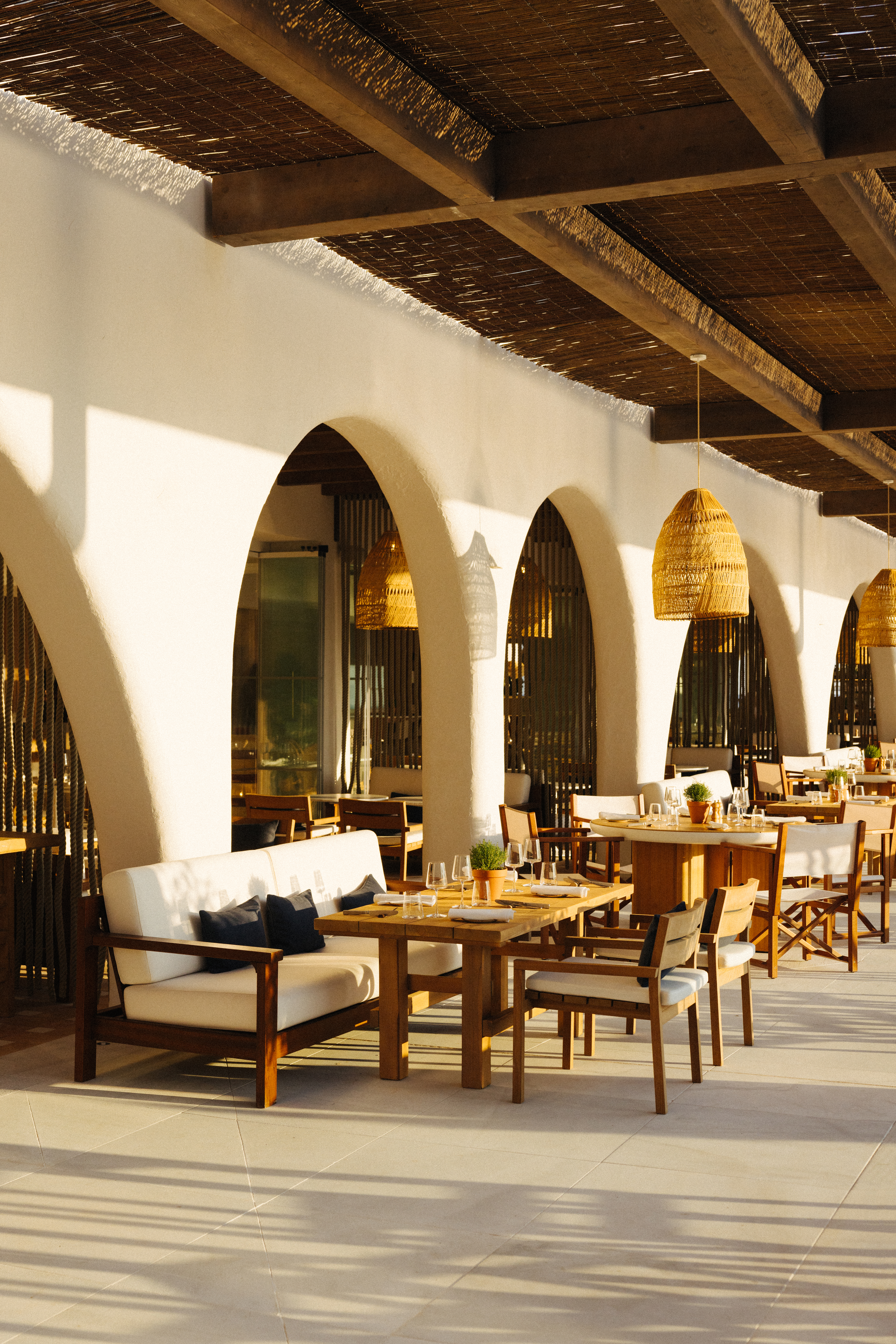 Sunny outdoor restaurant patio, white arches, wooden tables, woven ceiling.