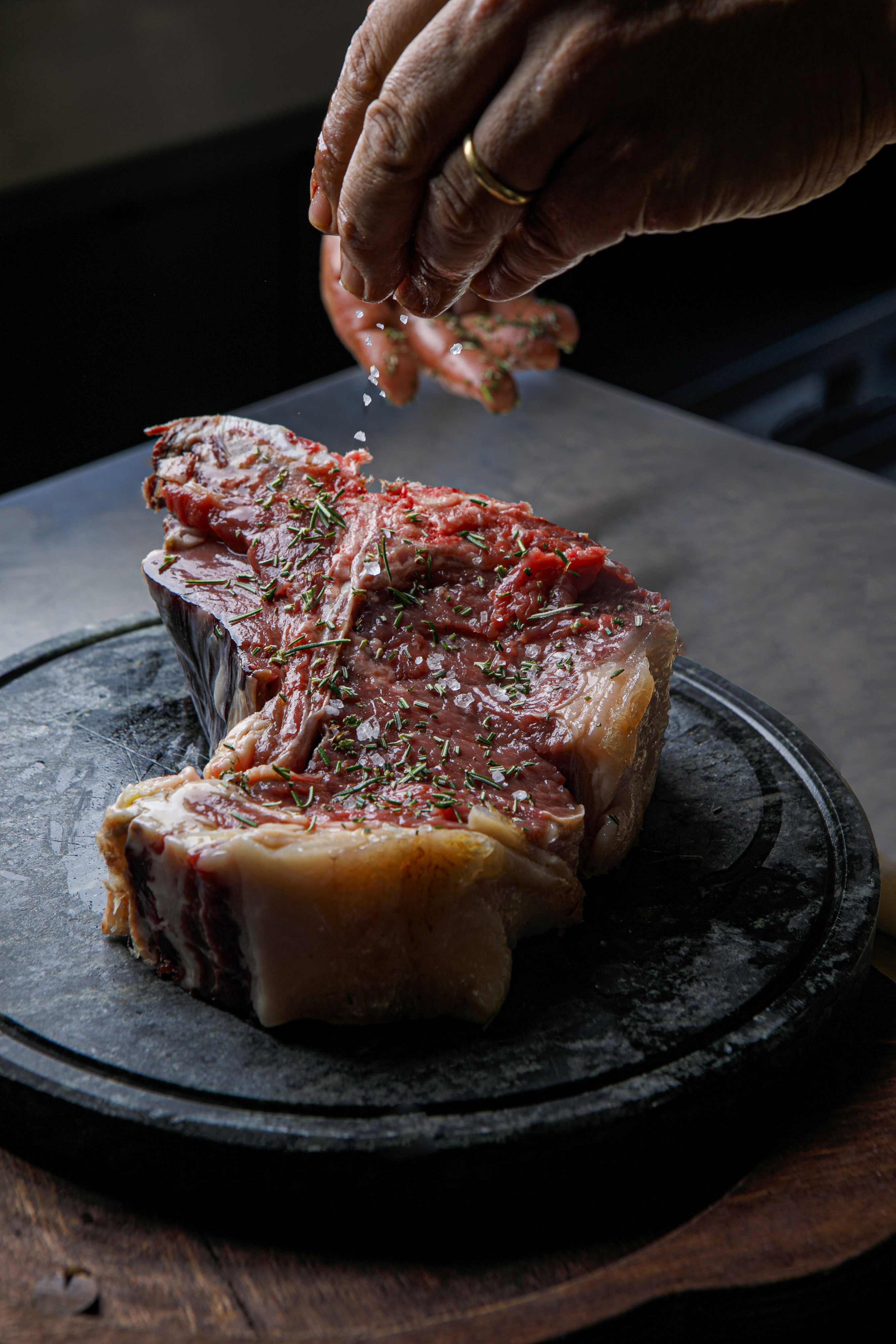 Chef's hand seasoning raw T-bone steak with coarse salt and rosemary.