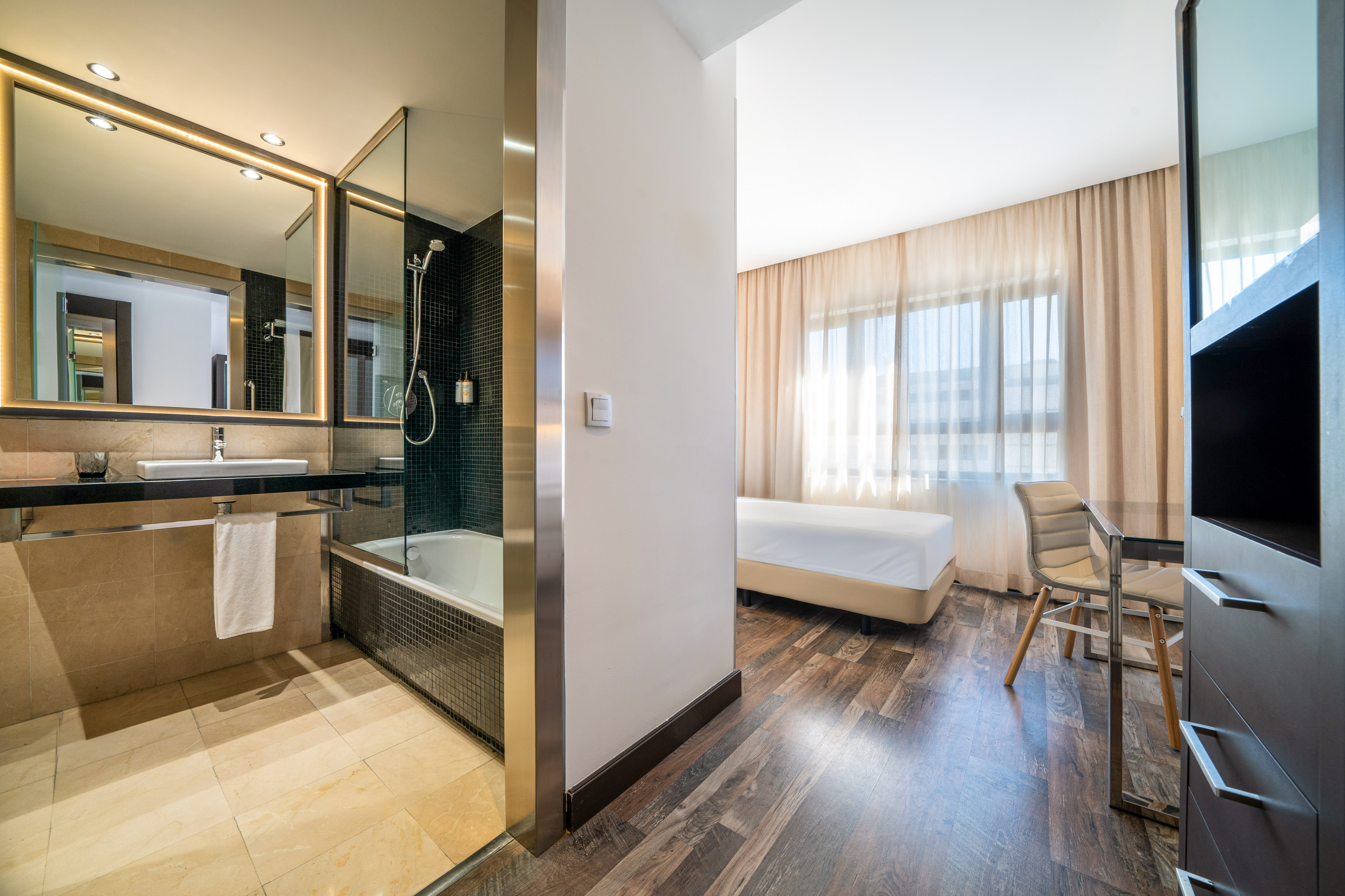 Modern hotel room with sleek bathroom, tub, vanity, and bright bedroom.