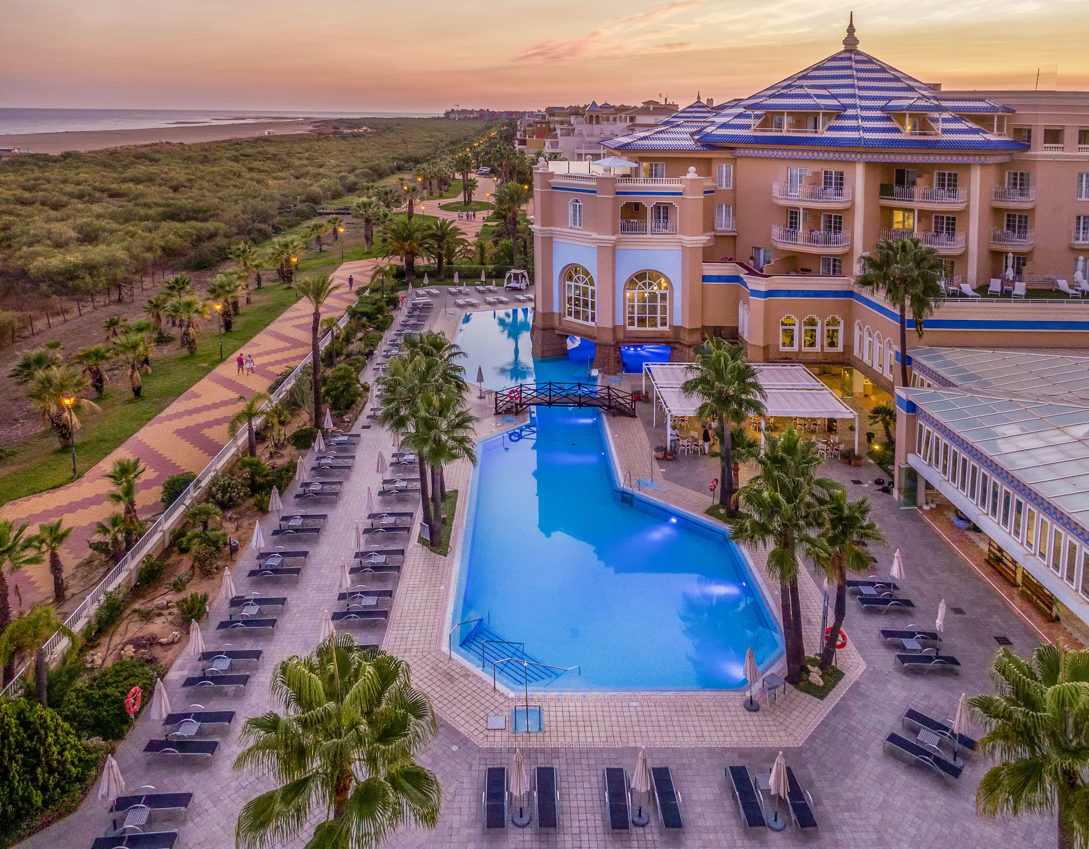 Aerial view of a luxury beach resort with blue pools, palm trees, and sunset sky.