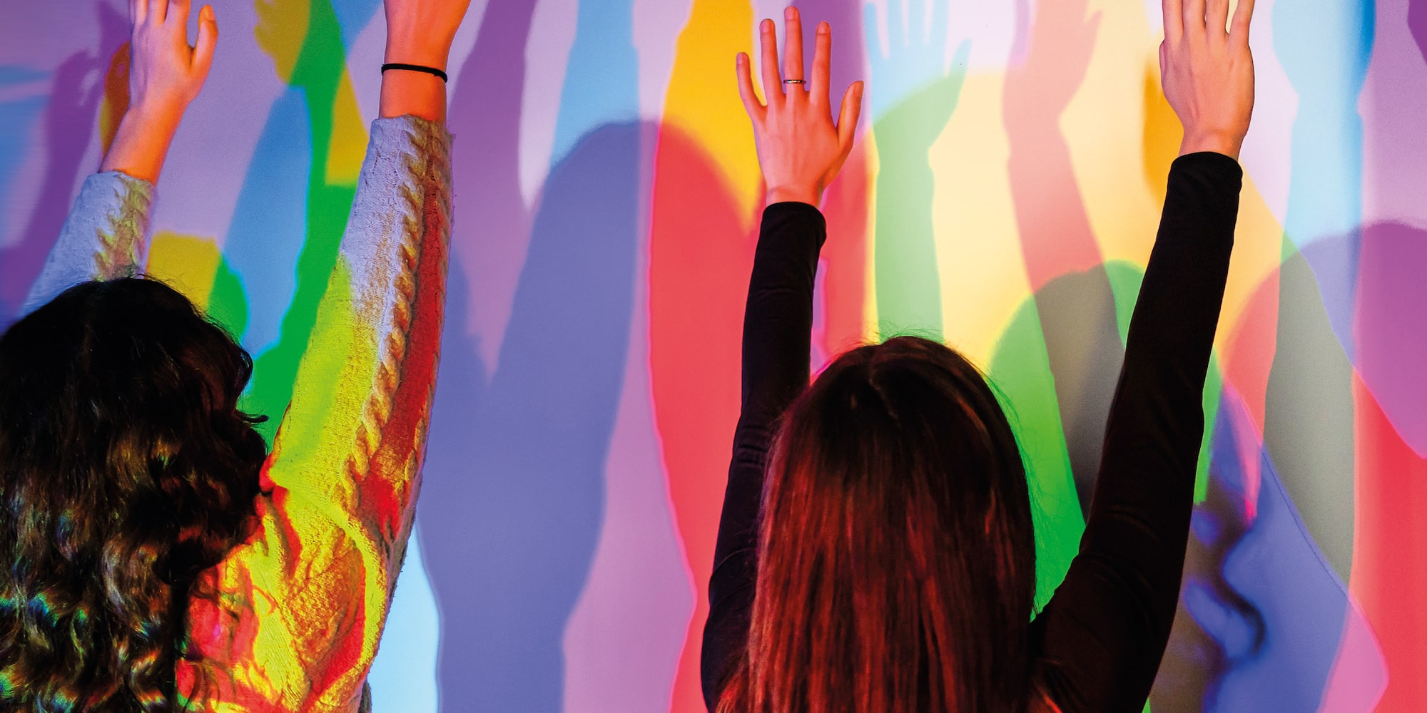 Two people raising hands, casting vibrant colorful shadows on a wall.