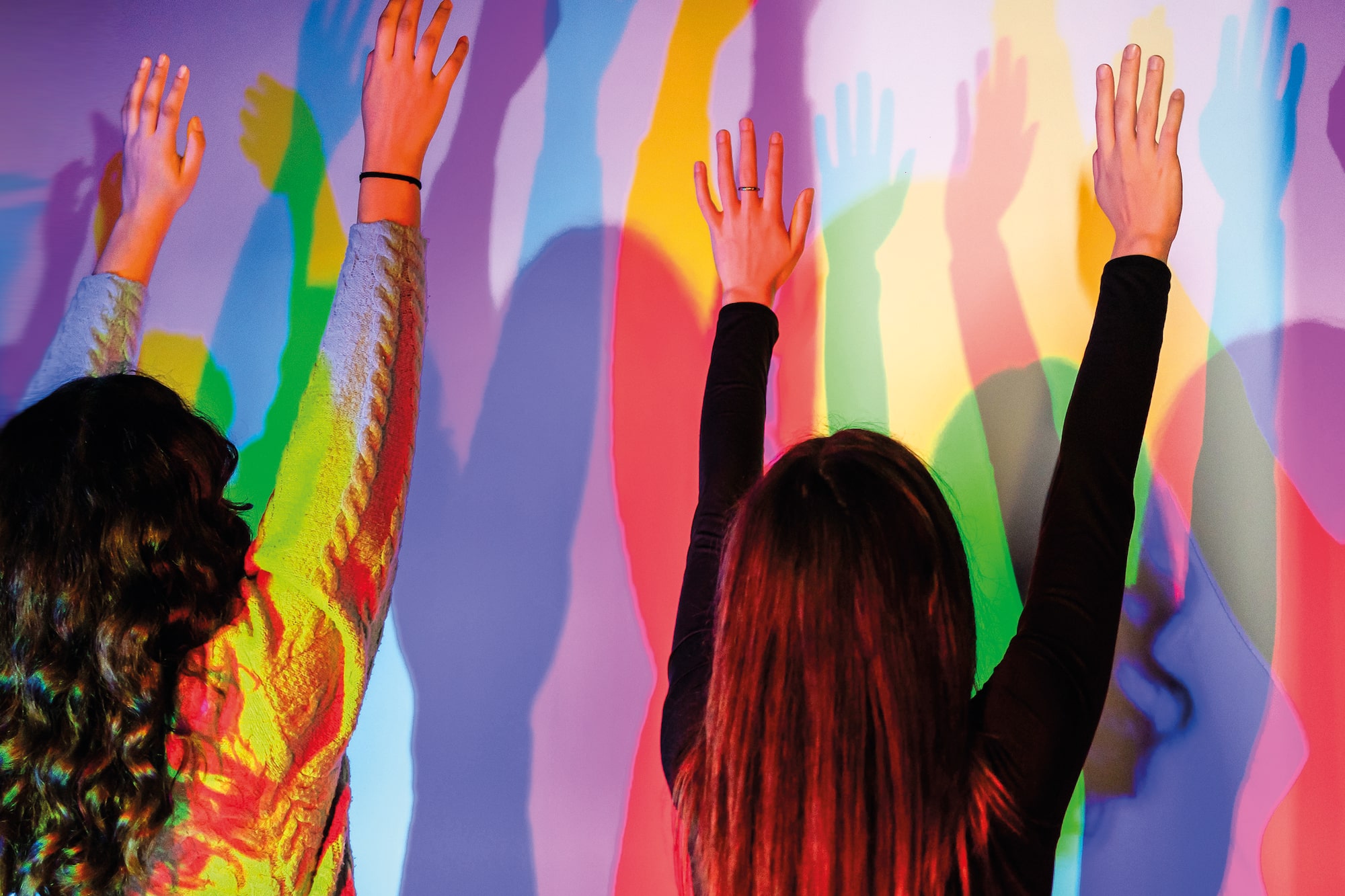 Two people raising hands, casting vibrant colorful shadows on a wall.