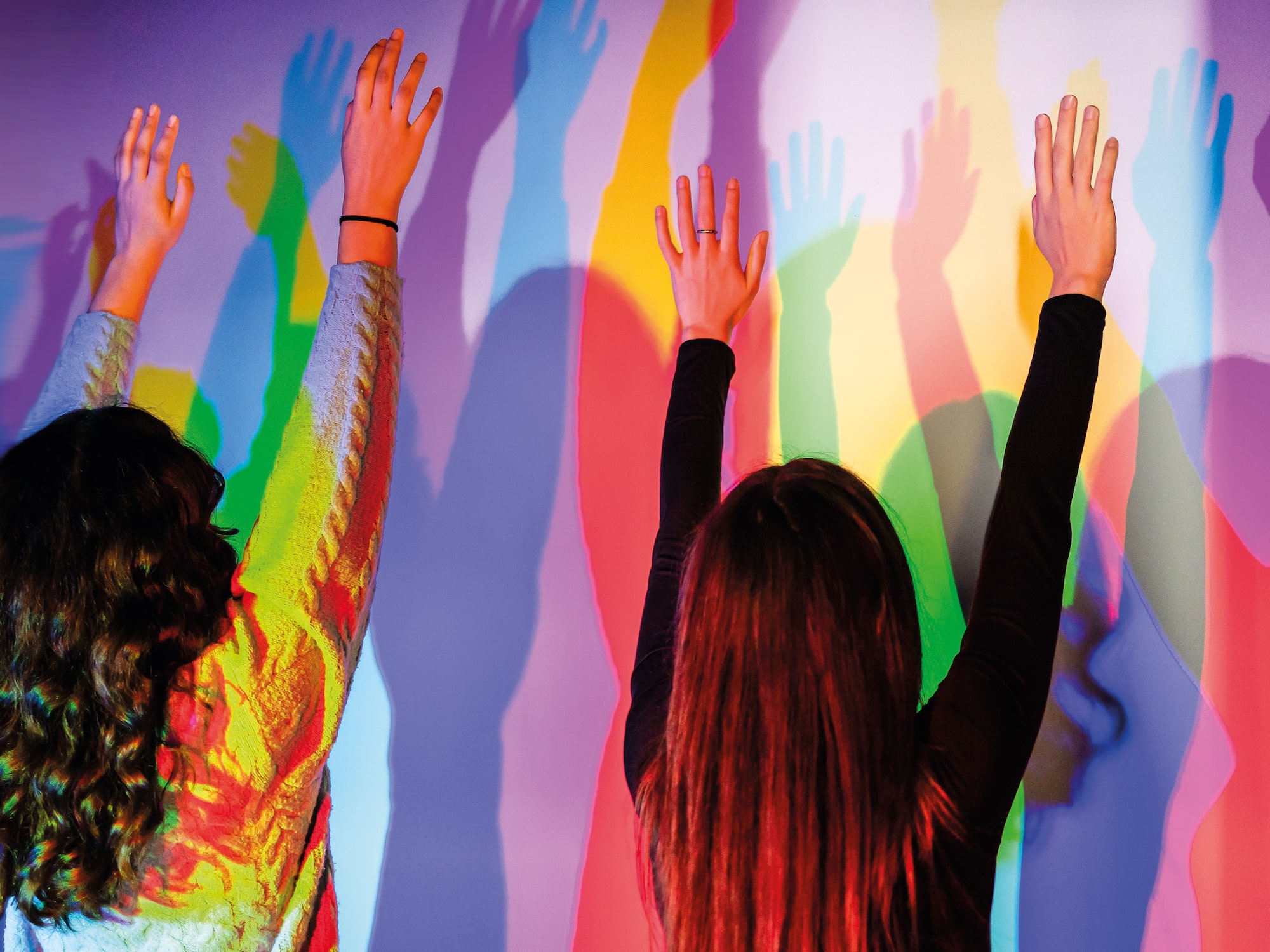 Two people raising hands, casting vibrant colorful shadows on a wall.