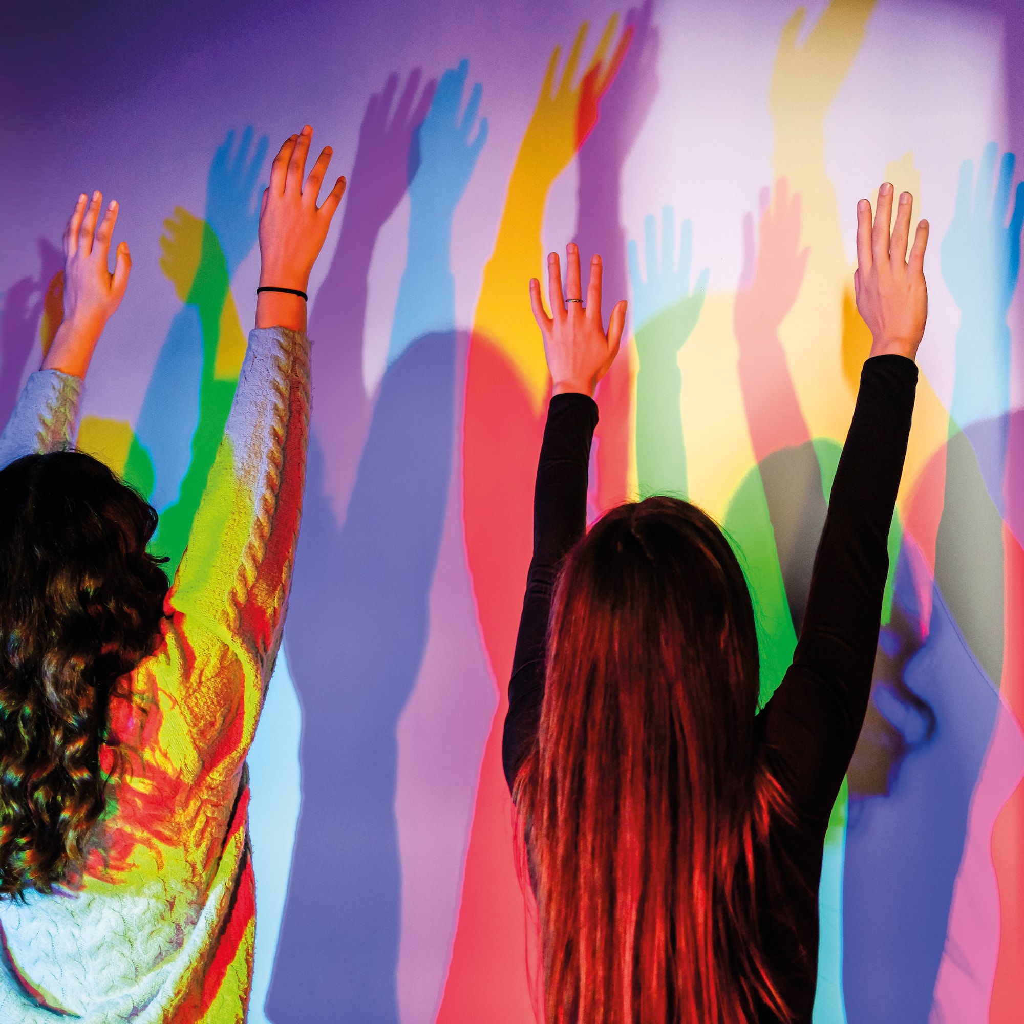 Two people raising hands, casting vibrant colorful shadows on a wall.