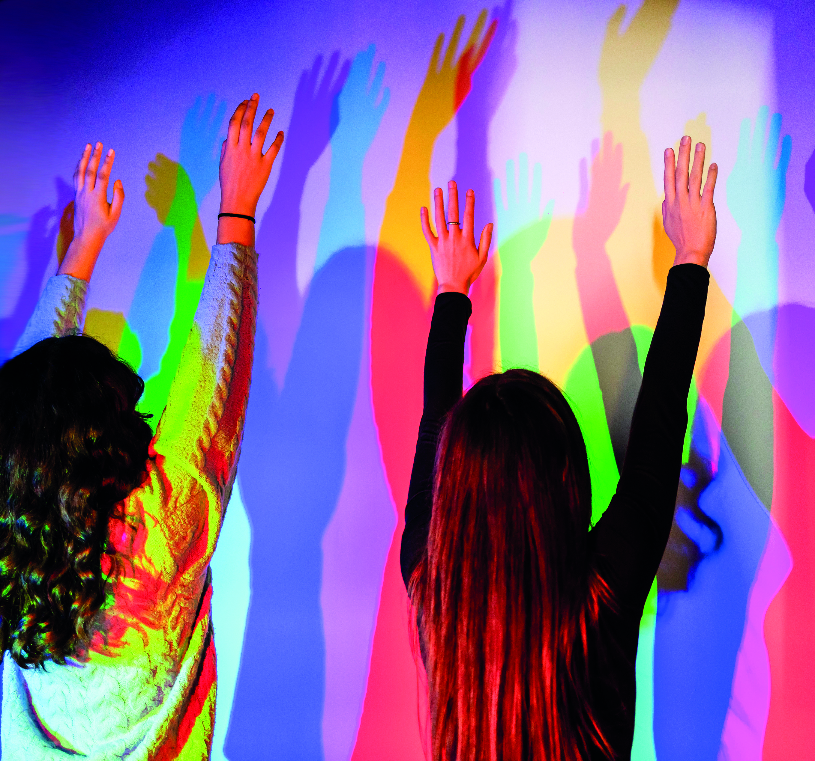 Two people raising hands, casting vibrant colorful shadows on a wall.