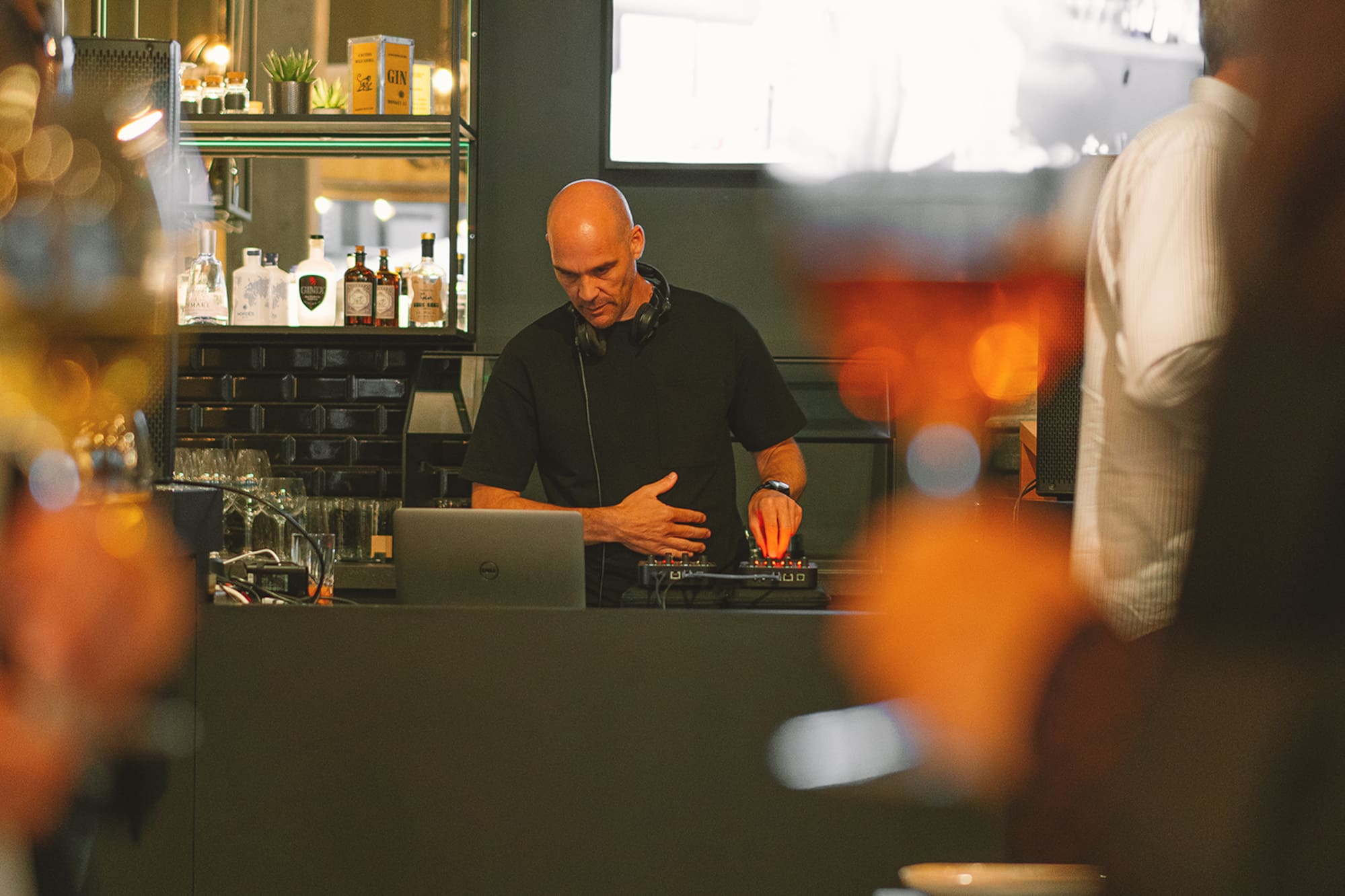 Bald DJ mixing music at a modern bar event, surrounded by bottles and glasses.