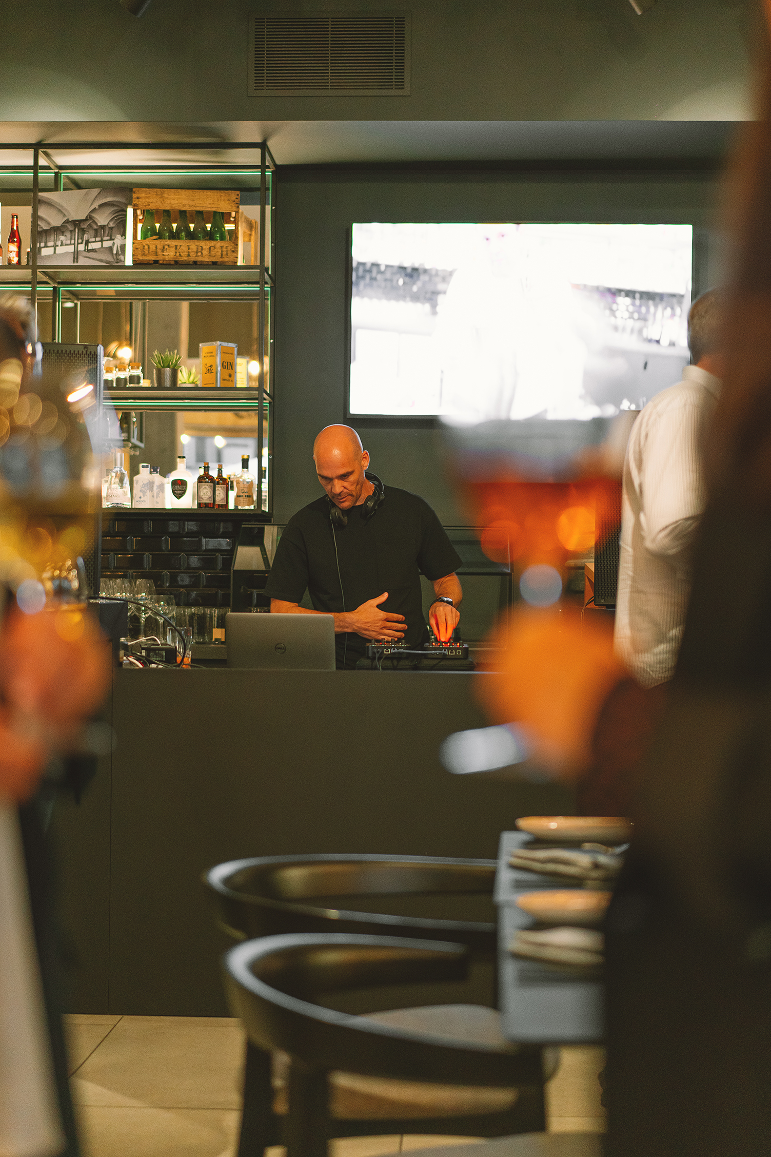 Bald DJ mixing music at a modern bar event, surrounded by bottles and glasses.