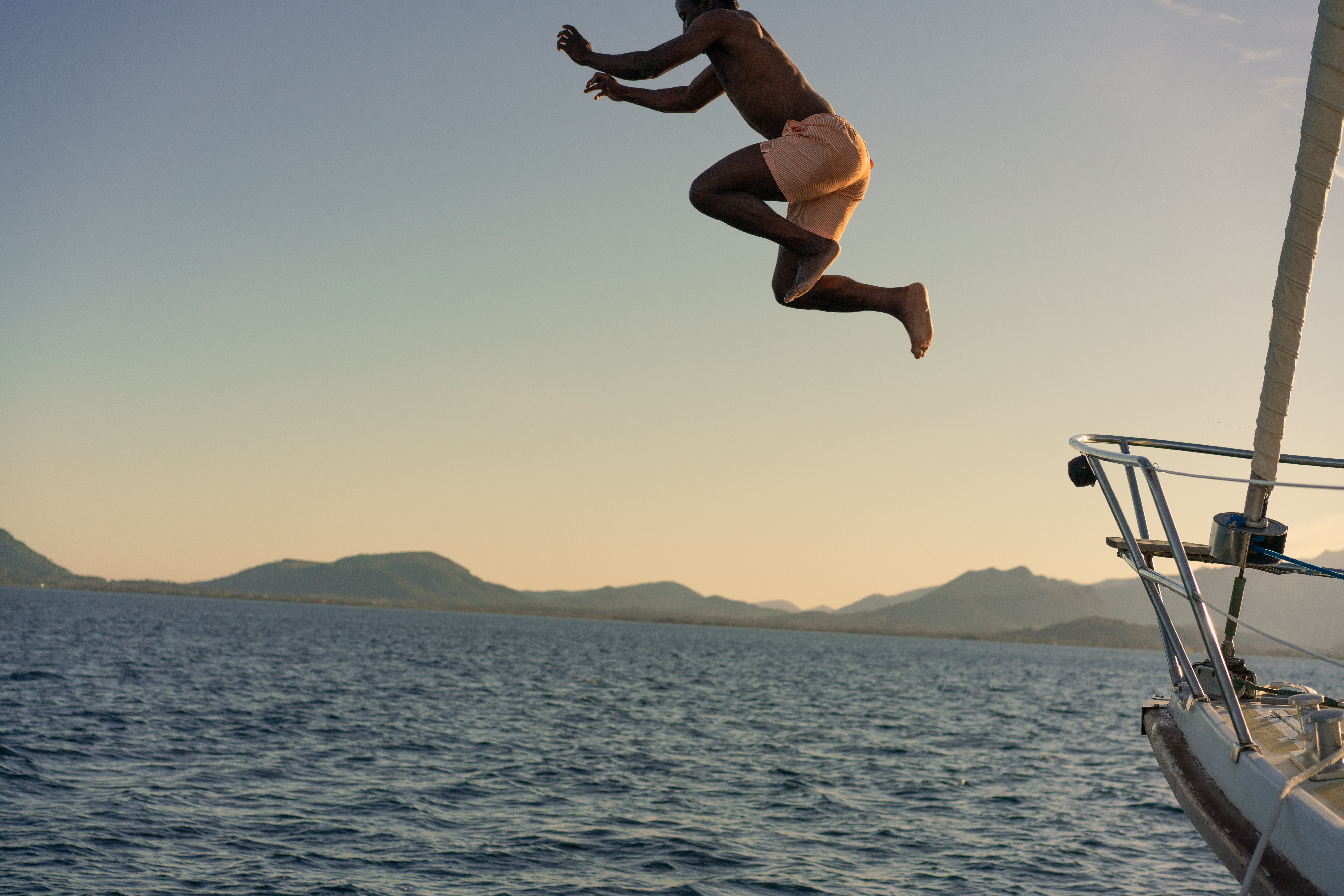 Man jumping from sailboat into ocean, distant mountains, gradient sky.