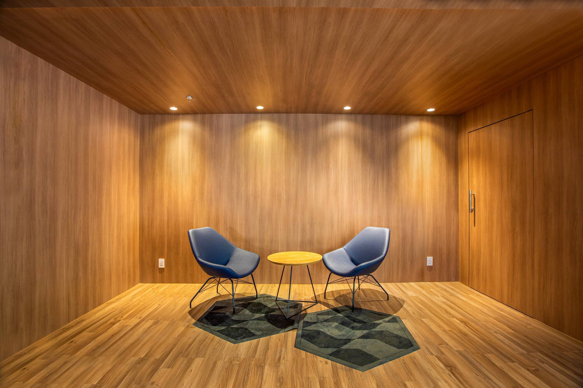 Wood-paneled lounge with two blue chairs, round table, and geometric rugs.