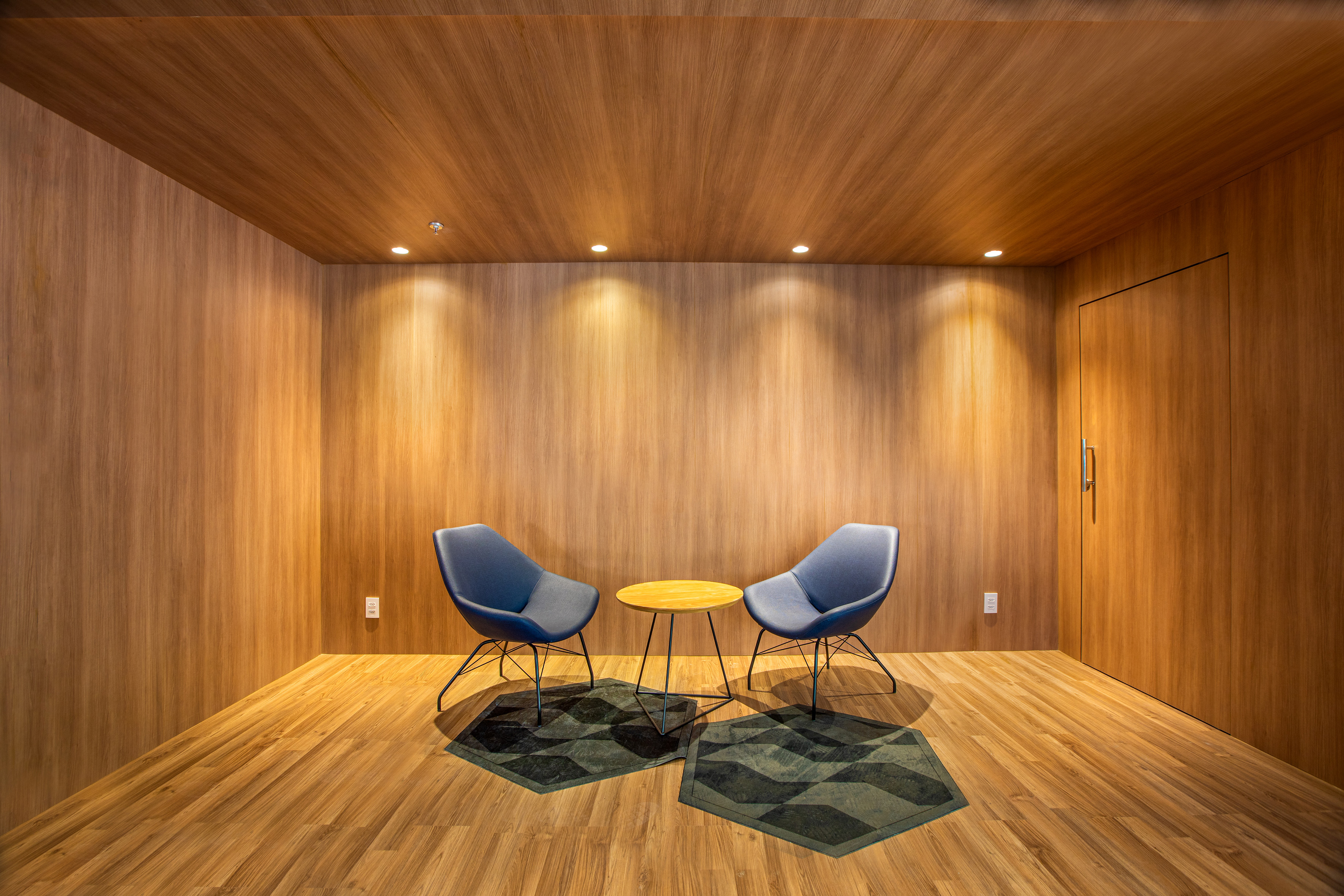 Wood-paneled lounge with two blue chairs, round table, and geometric rugs.