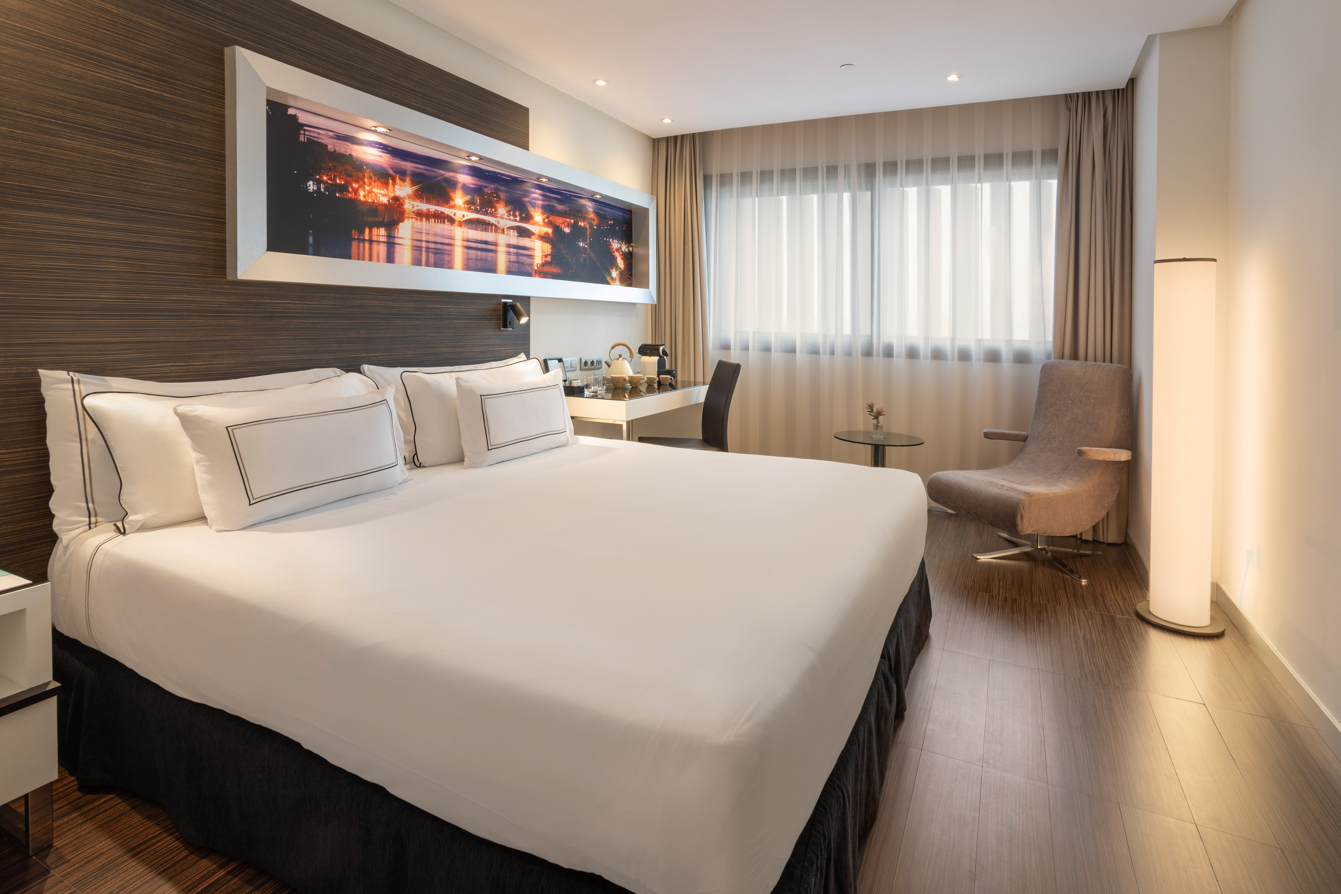 Modern hotel room with king bed, cityscape art, desk, and armchair.