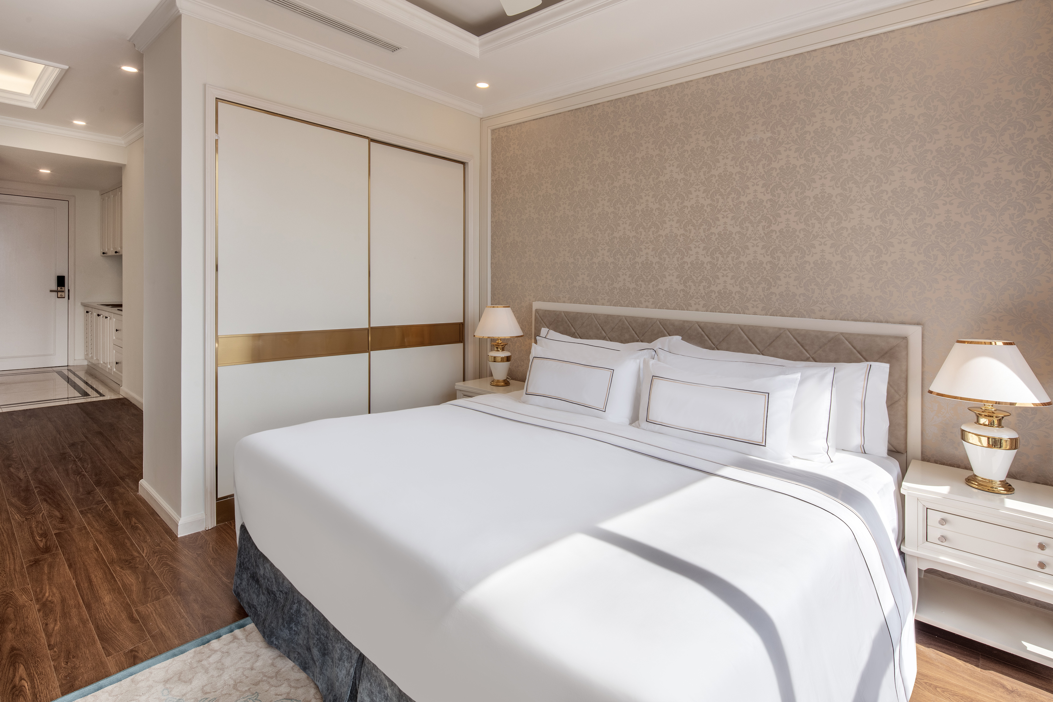 Elegant hotel bedroom with white bed, patterned wall, and gold closet doors.