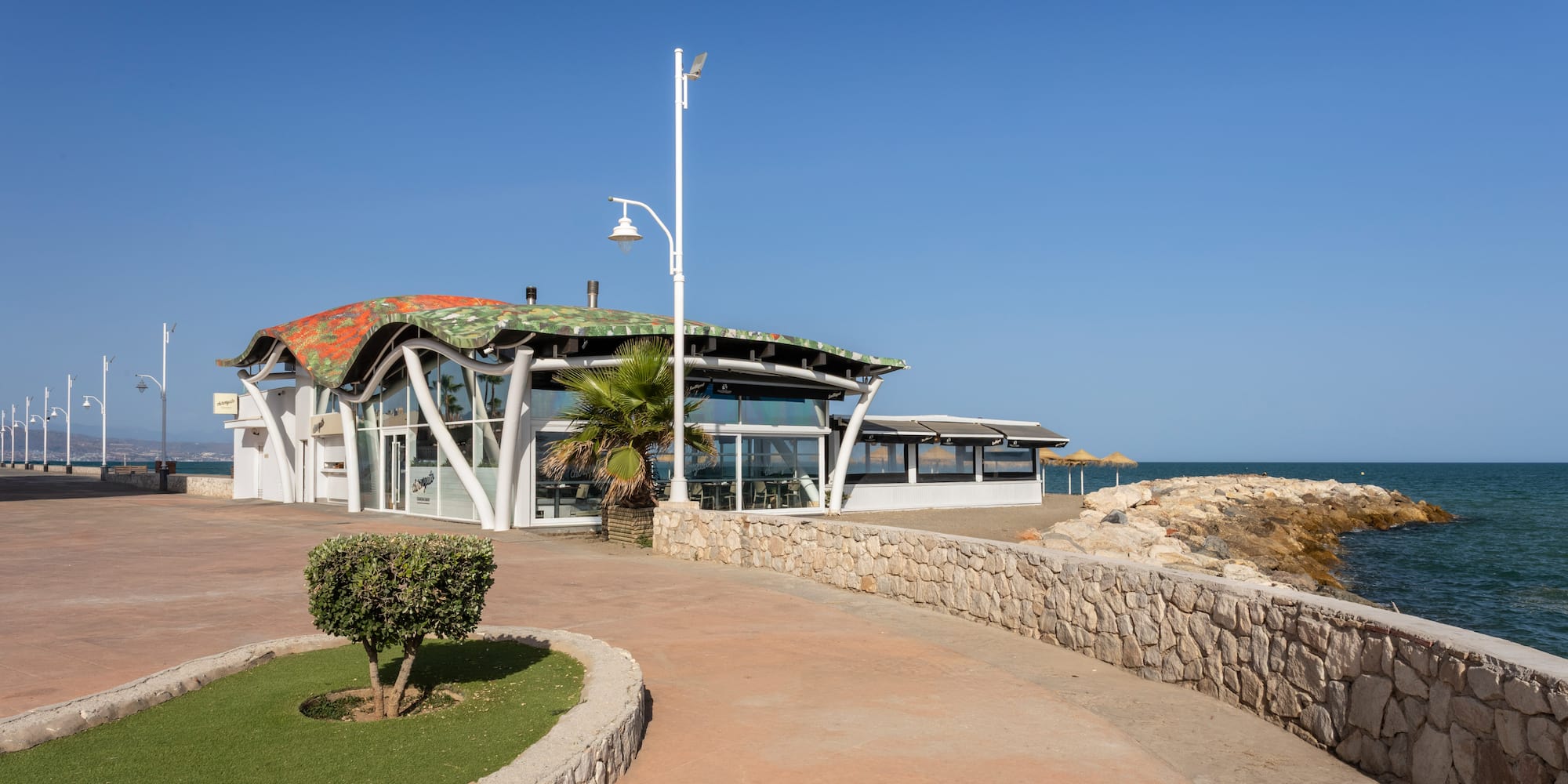 Modern beachfront restaurant with wavy roof, promenade, and blue sea.
