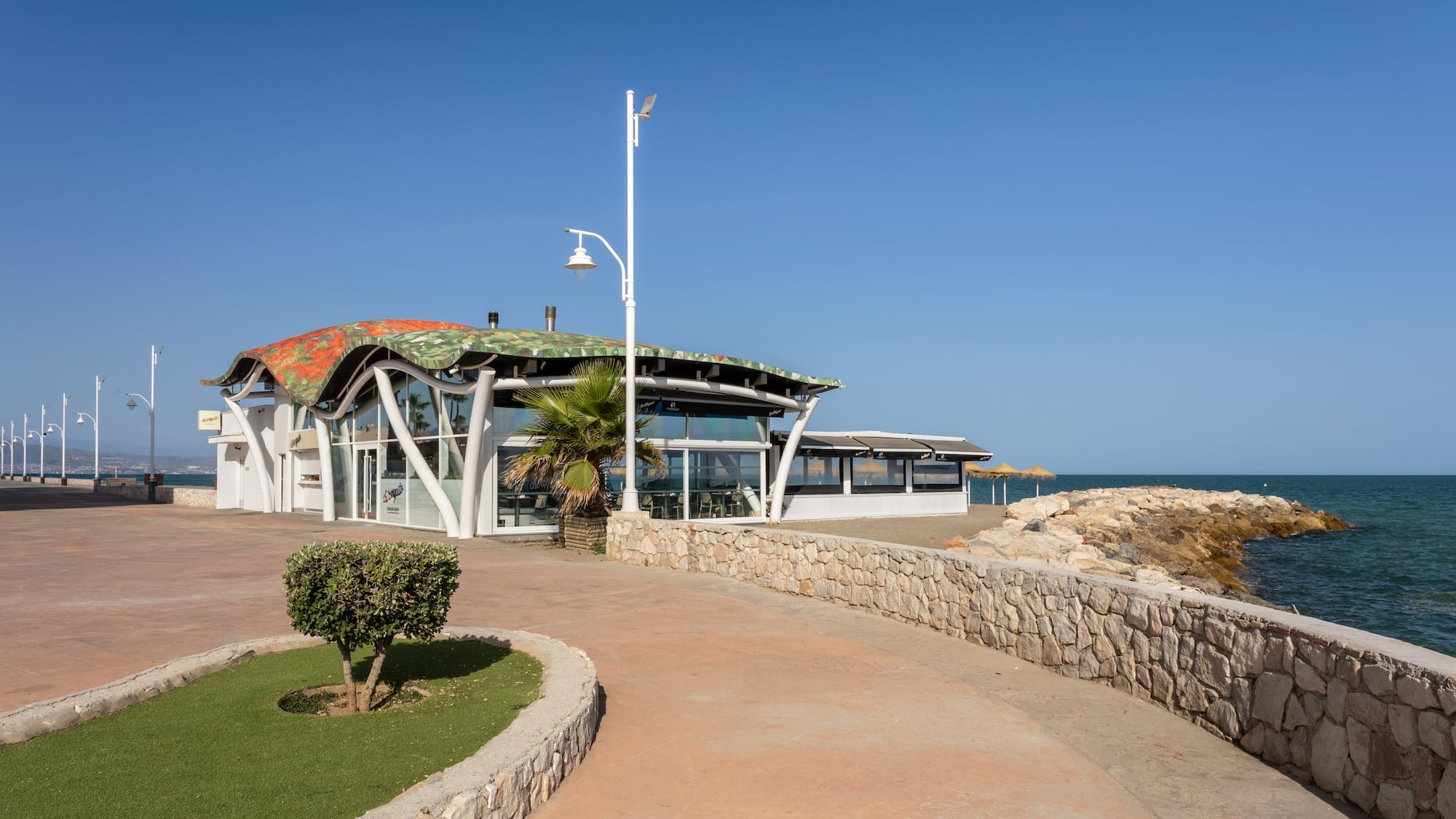 Modern beachfront restaurant with wavy roof, promenade, and blue sea.