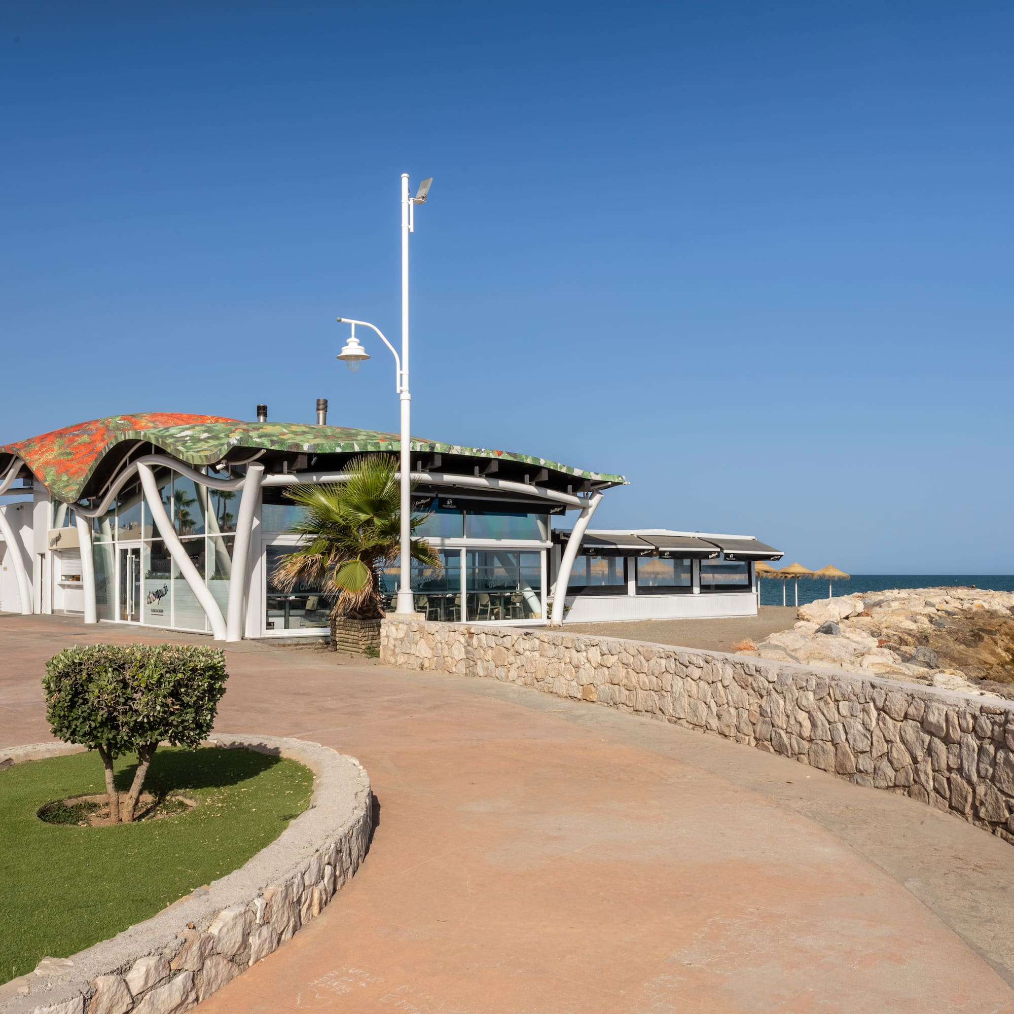 Modern beachfront restaurant with wavy roof, promenade, and blue sea.