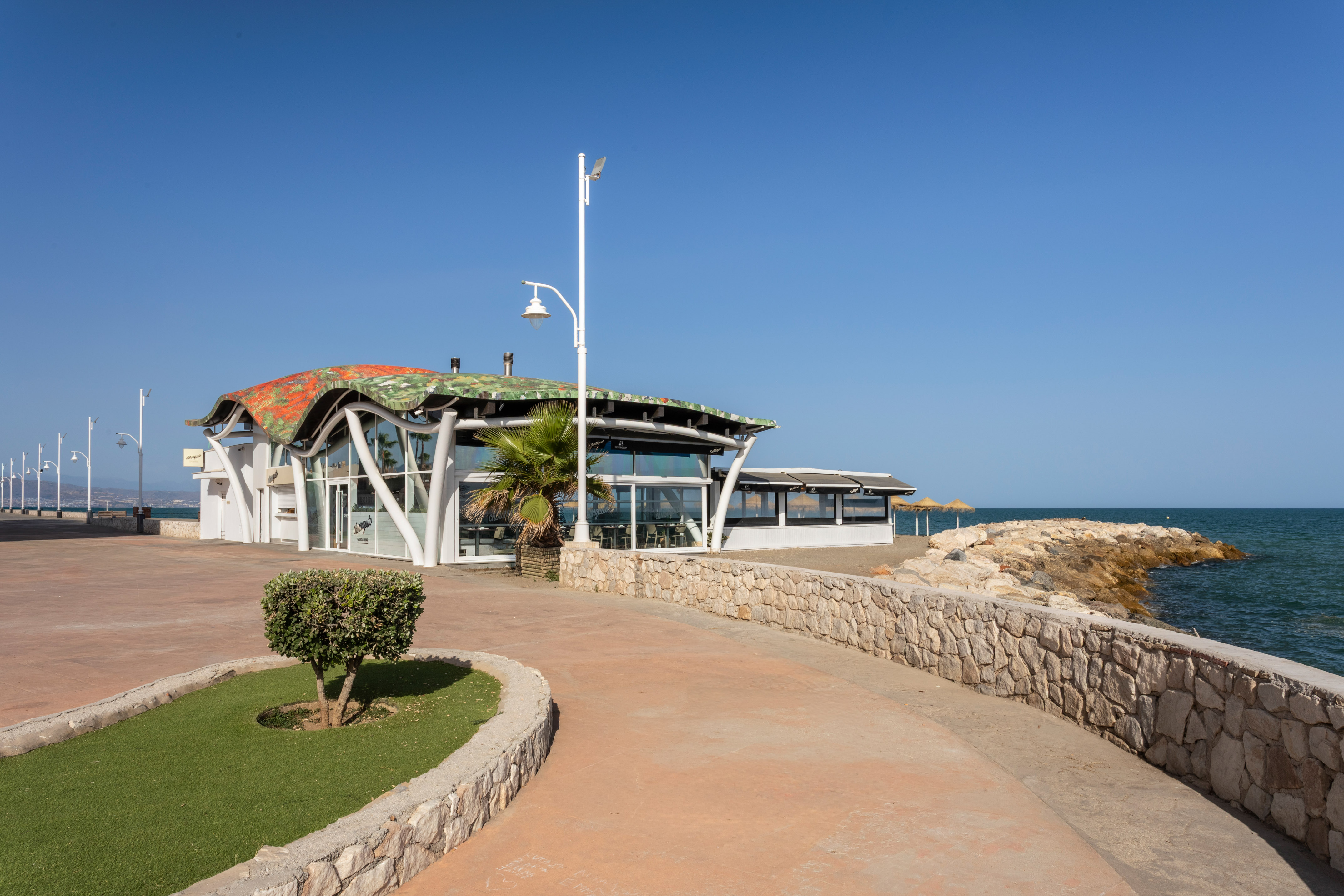 Modern beachfront restaurant with wavy roof, promenade, and blue sea.