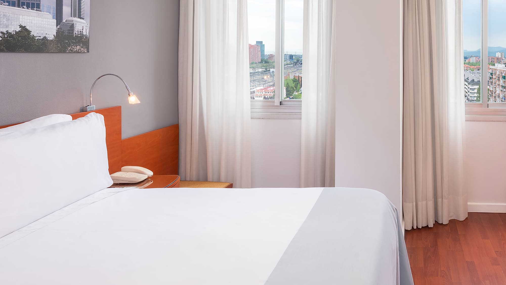Bright hotel room with city view, white bed, and wooden floor.