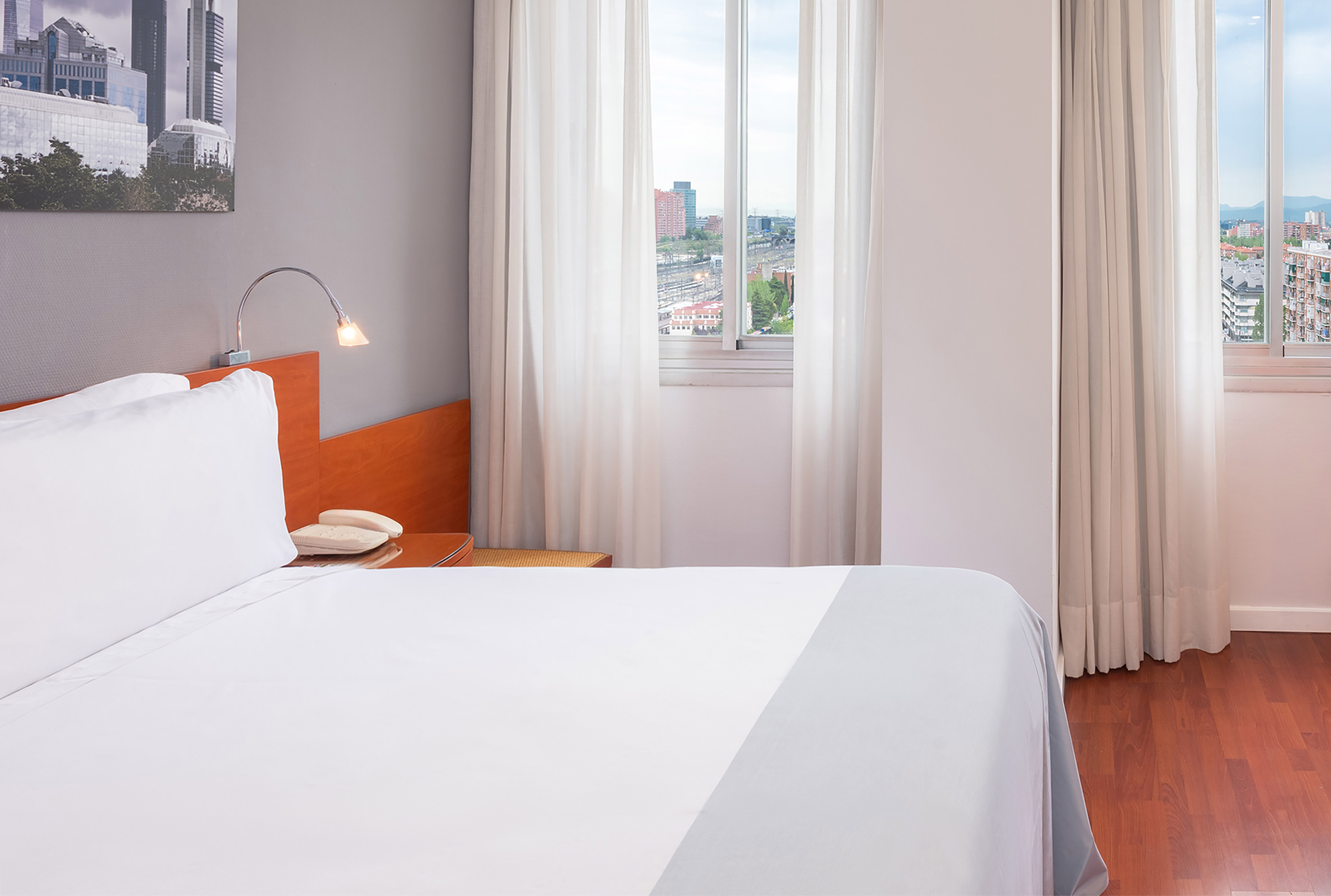 Bright hotel room with city view, white bed, and wooden floor.