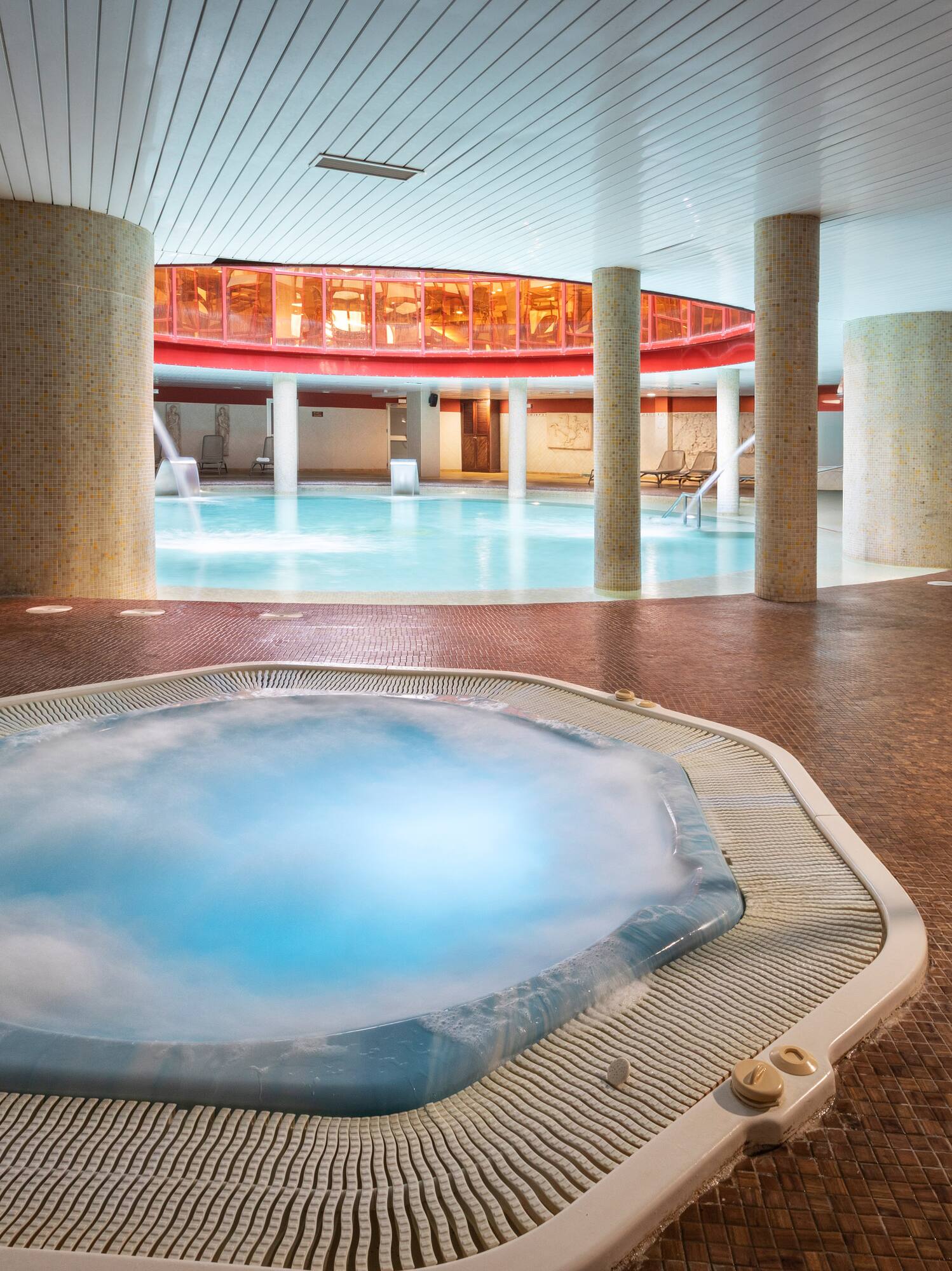 Gran piscina interior con jacuzzi y columnas en un spa moderno.