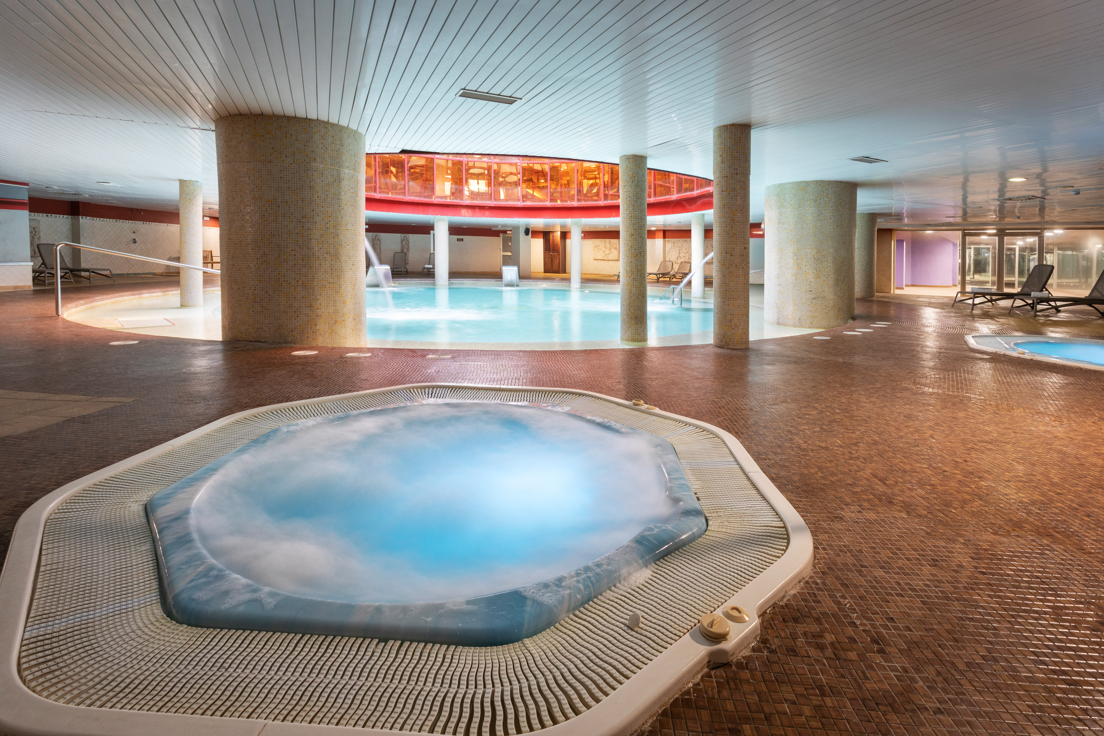 Gran piscina interior con jacuzzi y columnas en un spa moderno.