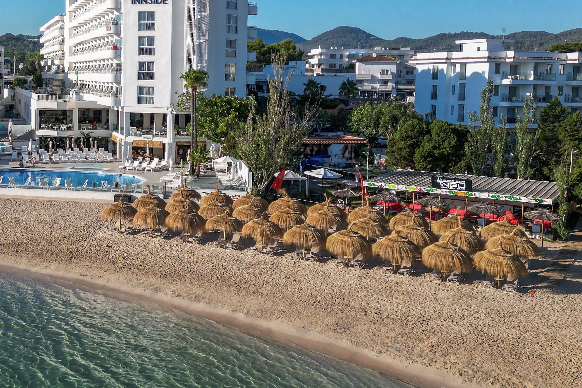 Aerial view of INNSIDE beach hotel, straw umbrellas, clear sea, and pool.