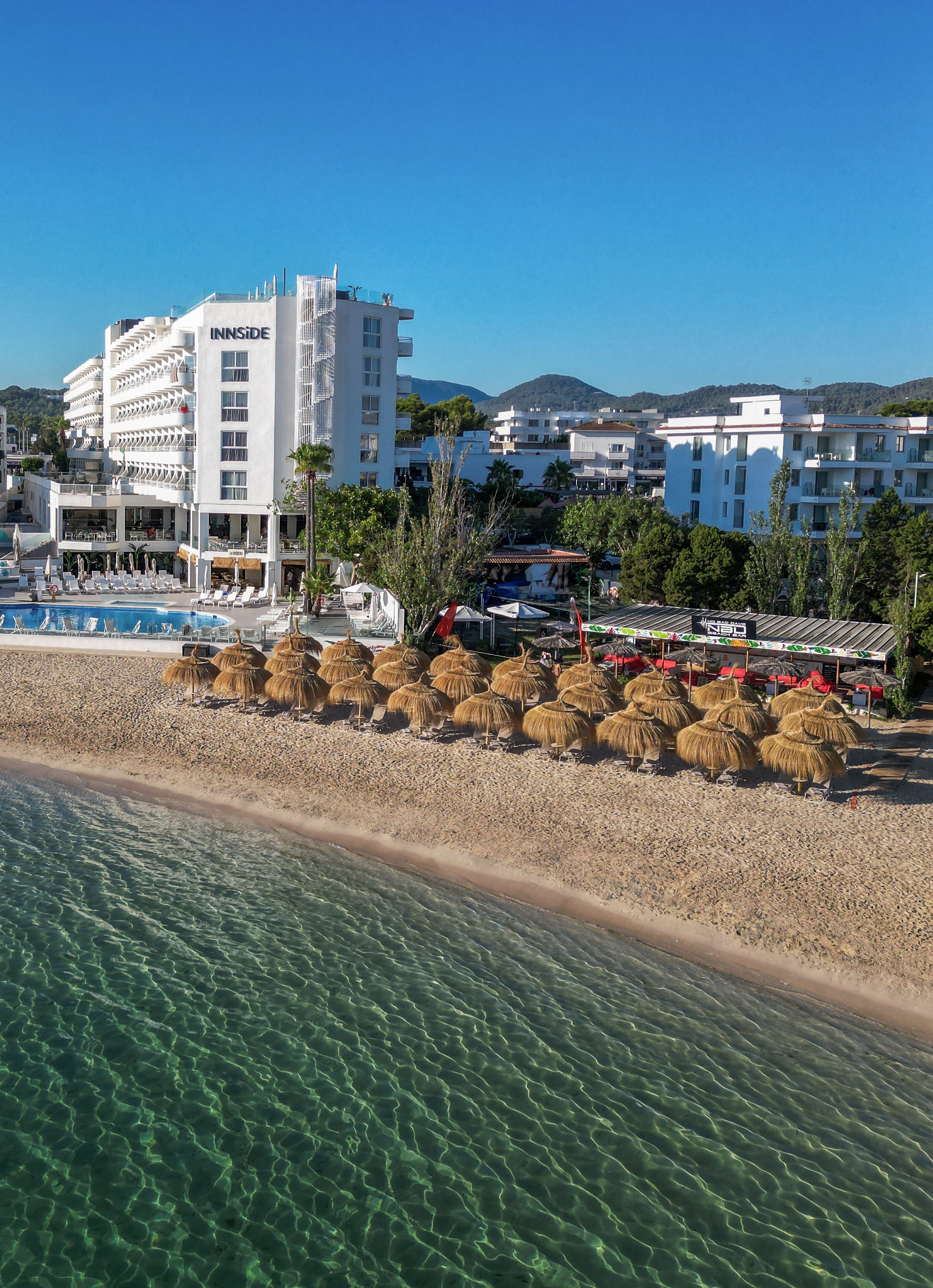 Aerial view of INNSIDE beach hotel, straw umbrellas, clear sea, and pool.