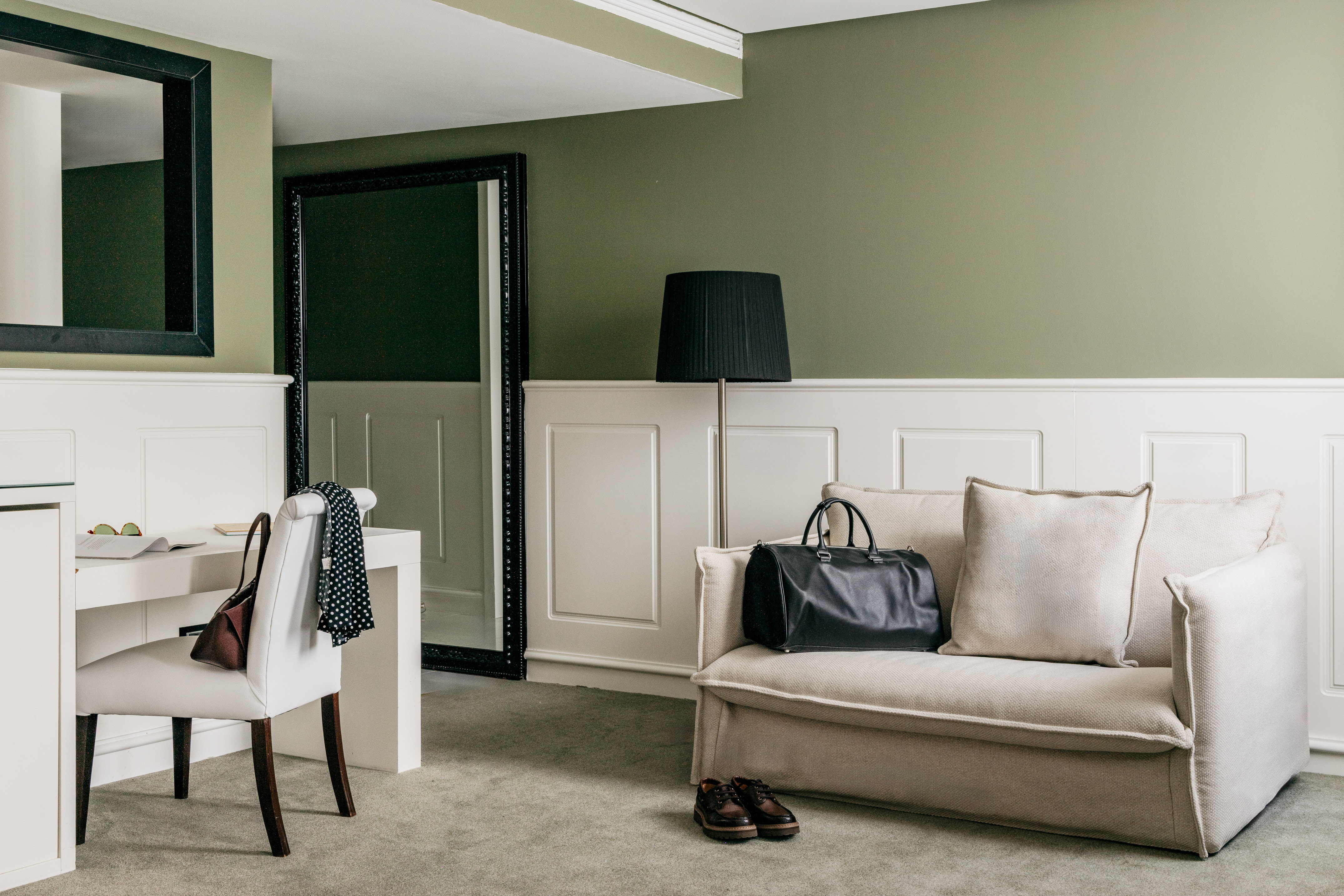 Olive green hotel room with beige sofa, black travel bag, desk, and mirrors.
