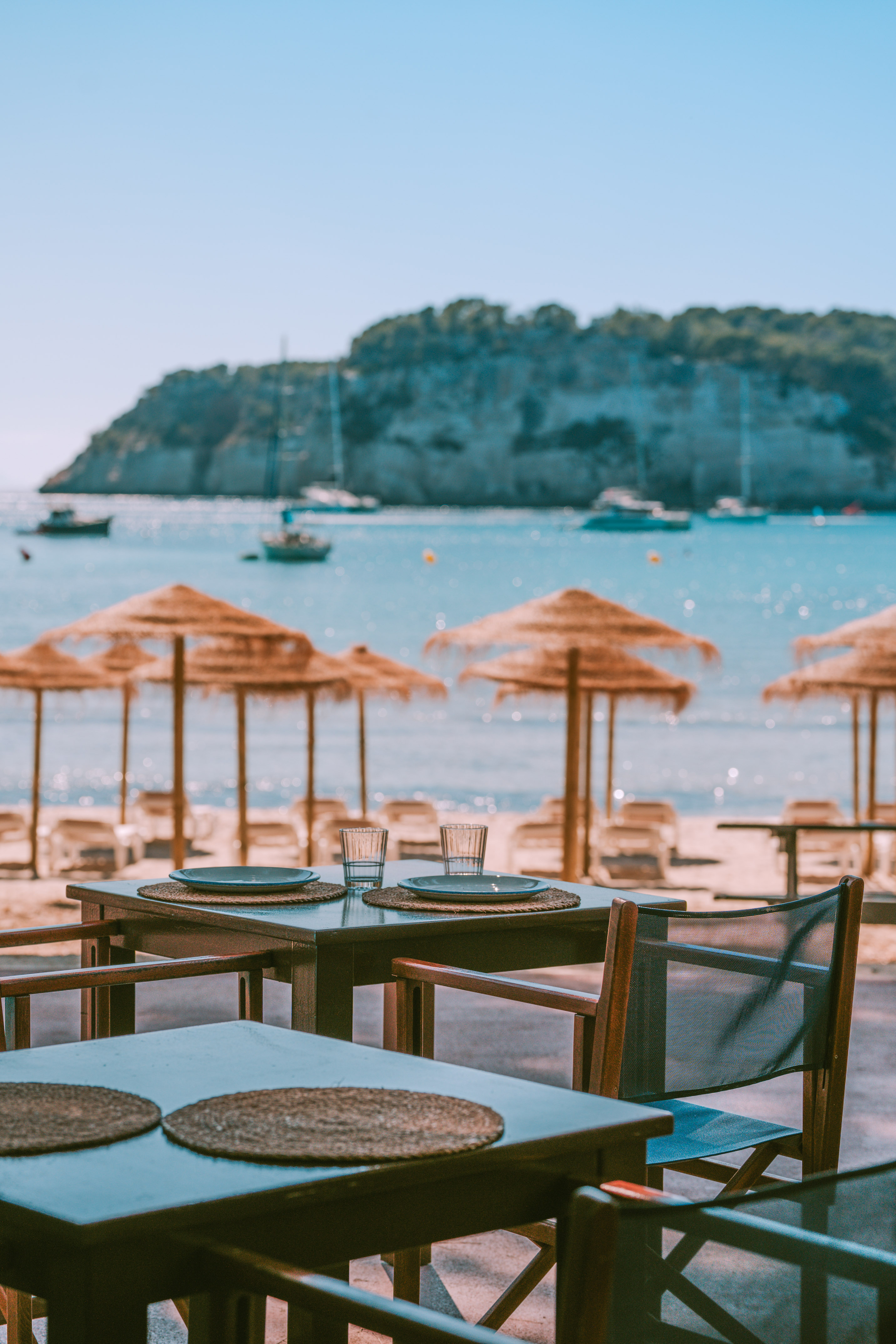 Beach restaurant tables, straw umbrellas, ocean with boats, island background.