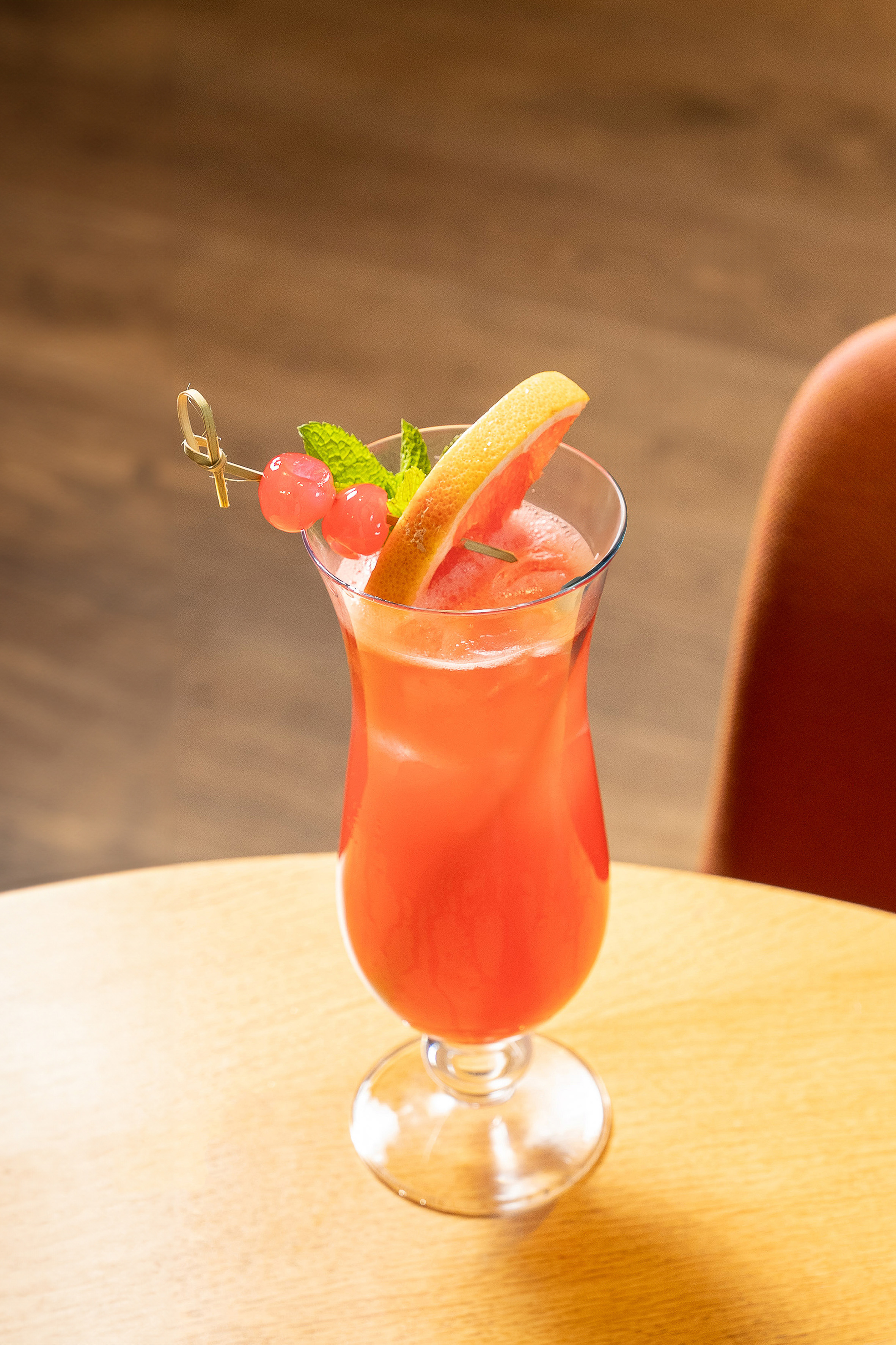 Red cocktail with grapefruit, mint, and cherries on a wooden table.