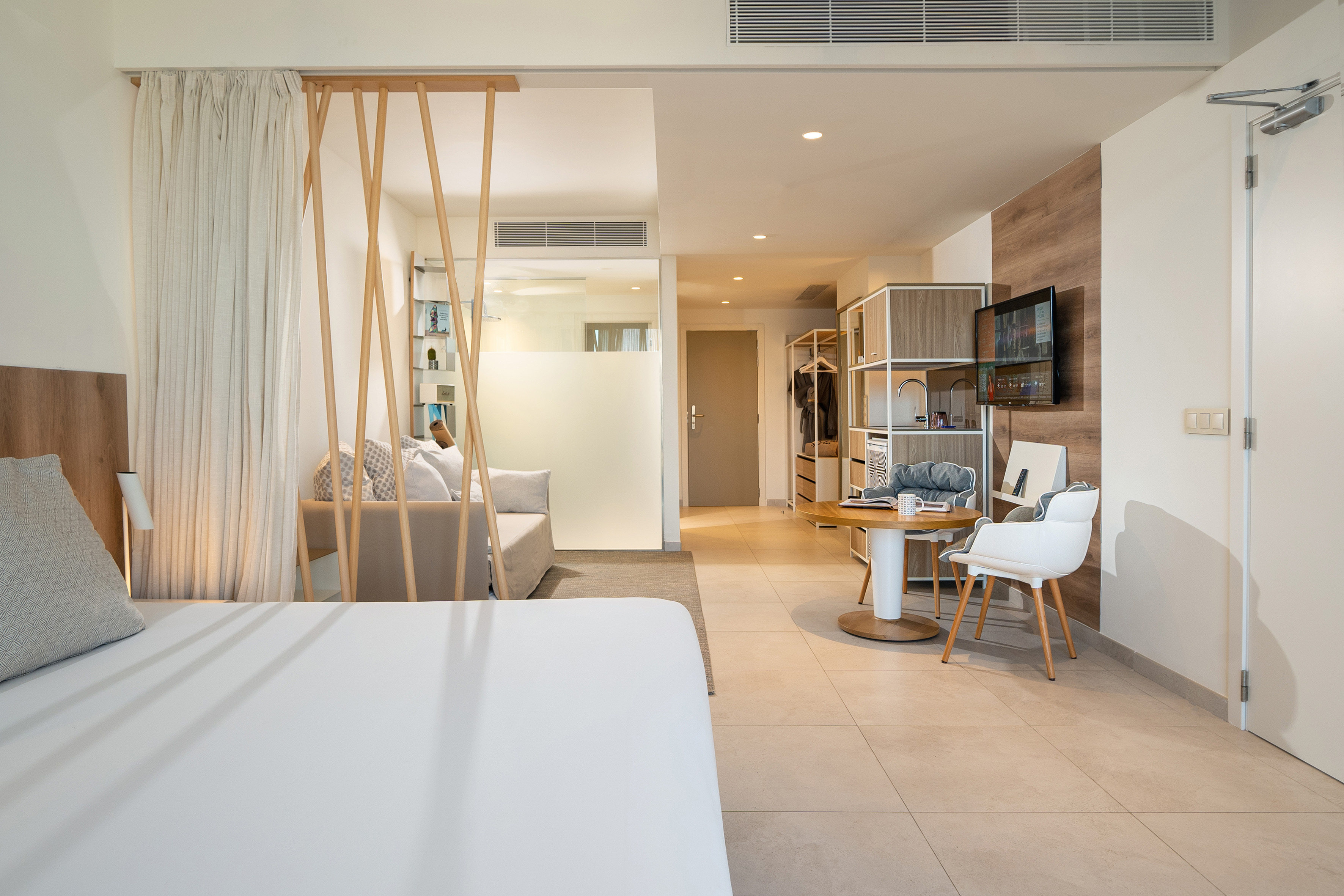 Modern hotel studio apartment with white bed, lounge area, TV, and wood accents.
