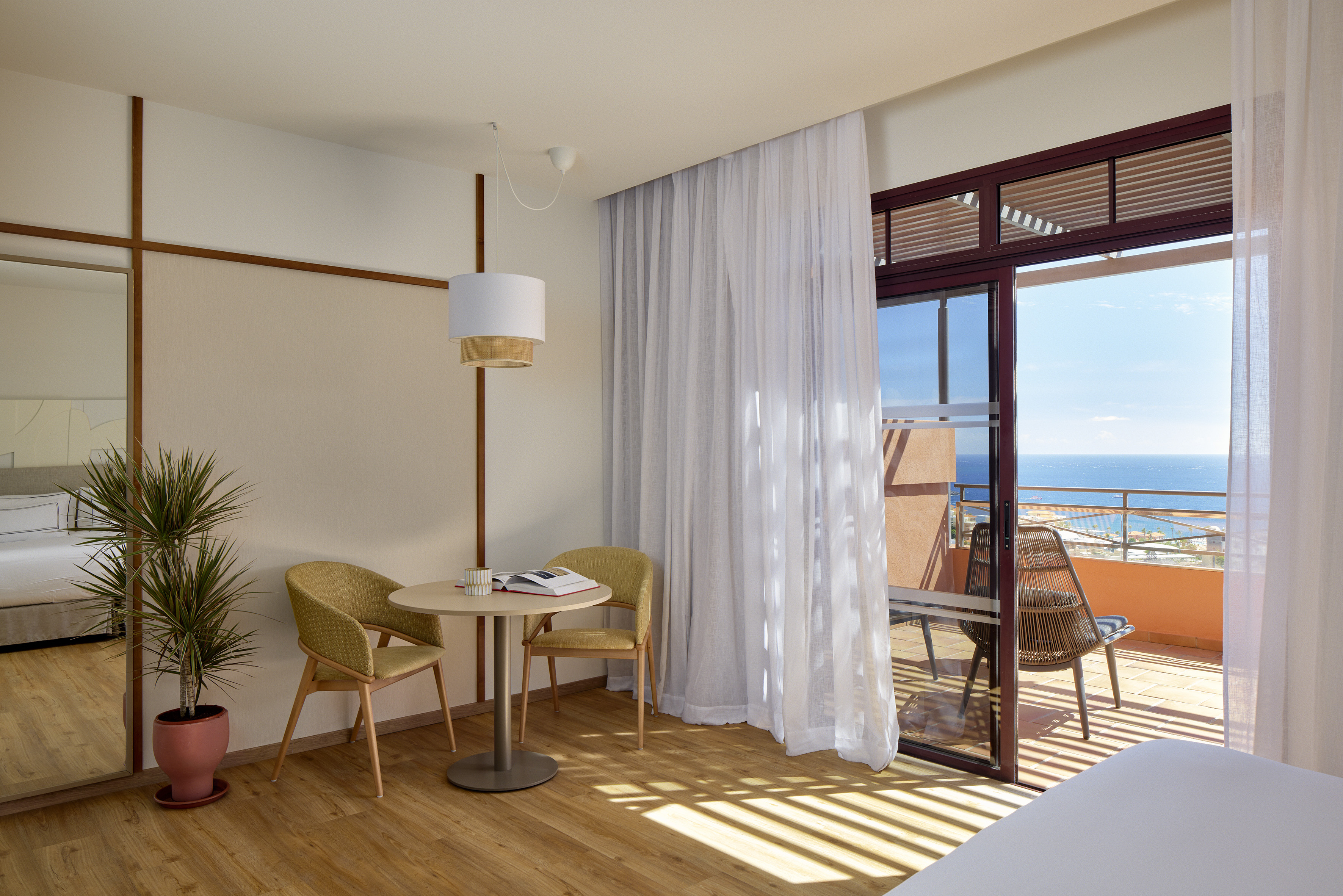 Sunny hotel room with ocean view balcony, wooden floor, and two chairs.