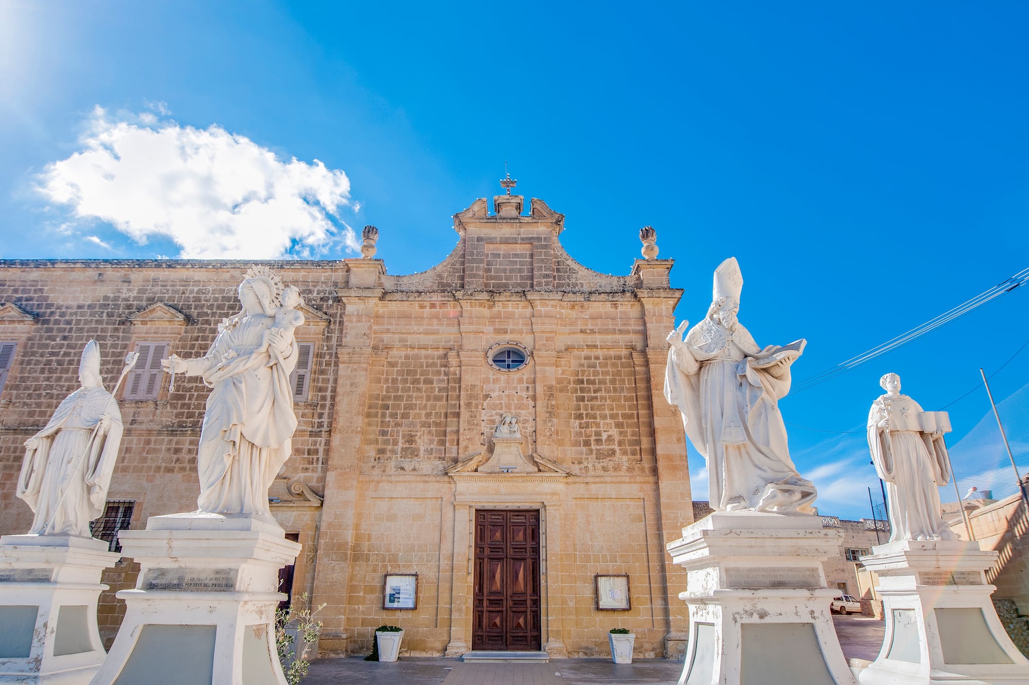 Historic stone church with statues of saints and Virgin Mary, bright blue sky.