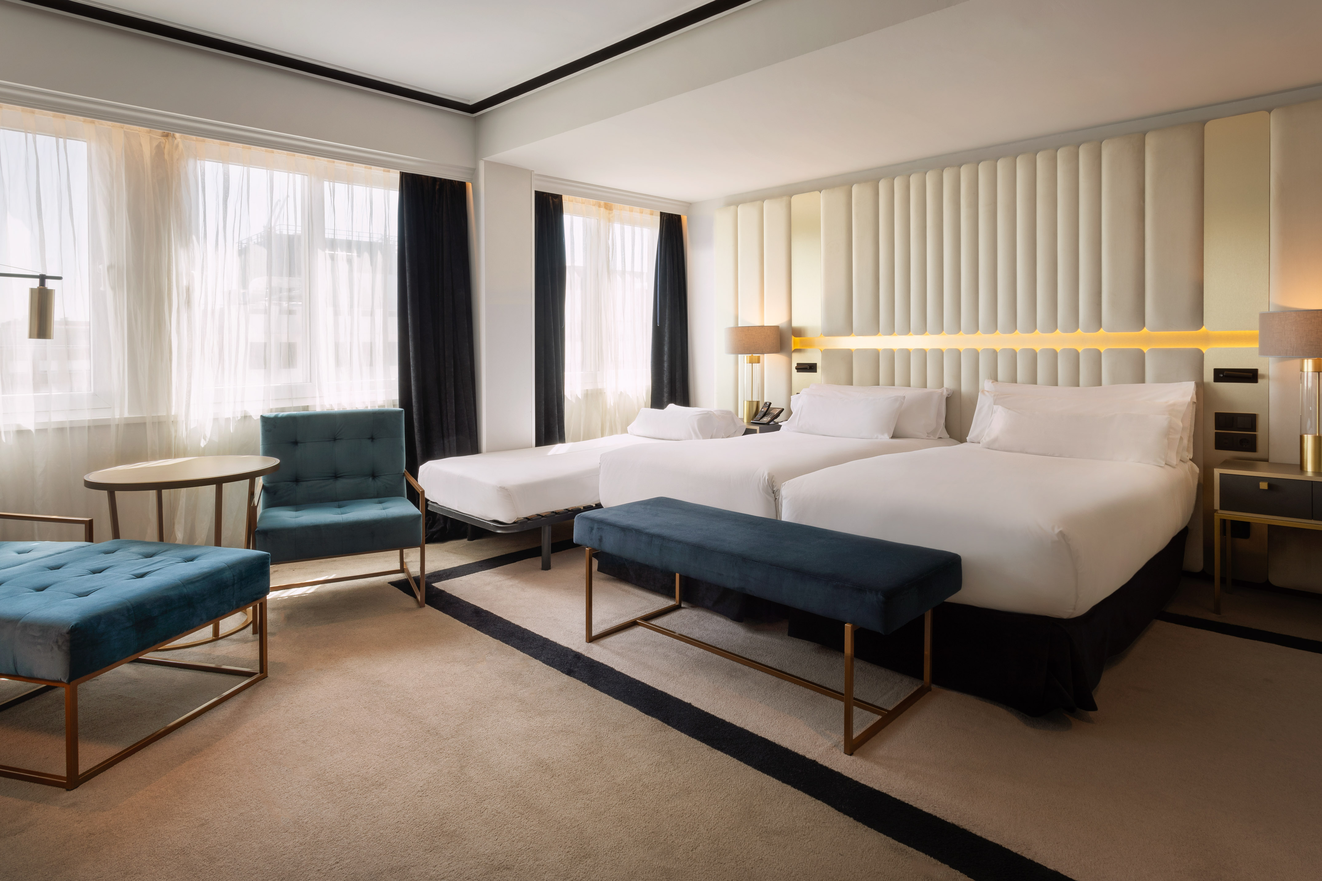 Modern hotel room with three beds, comfortable velvet seating, and bright windows.