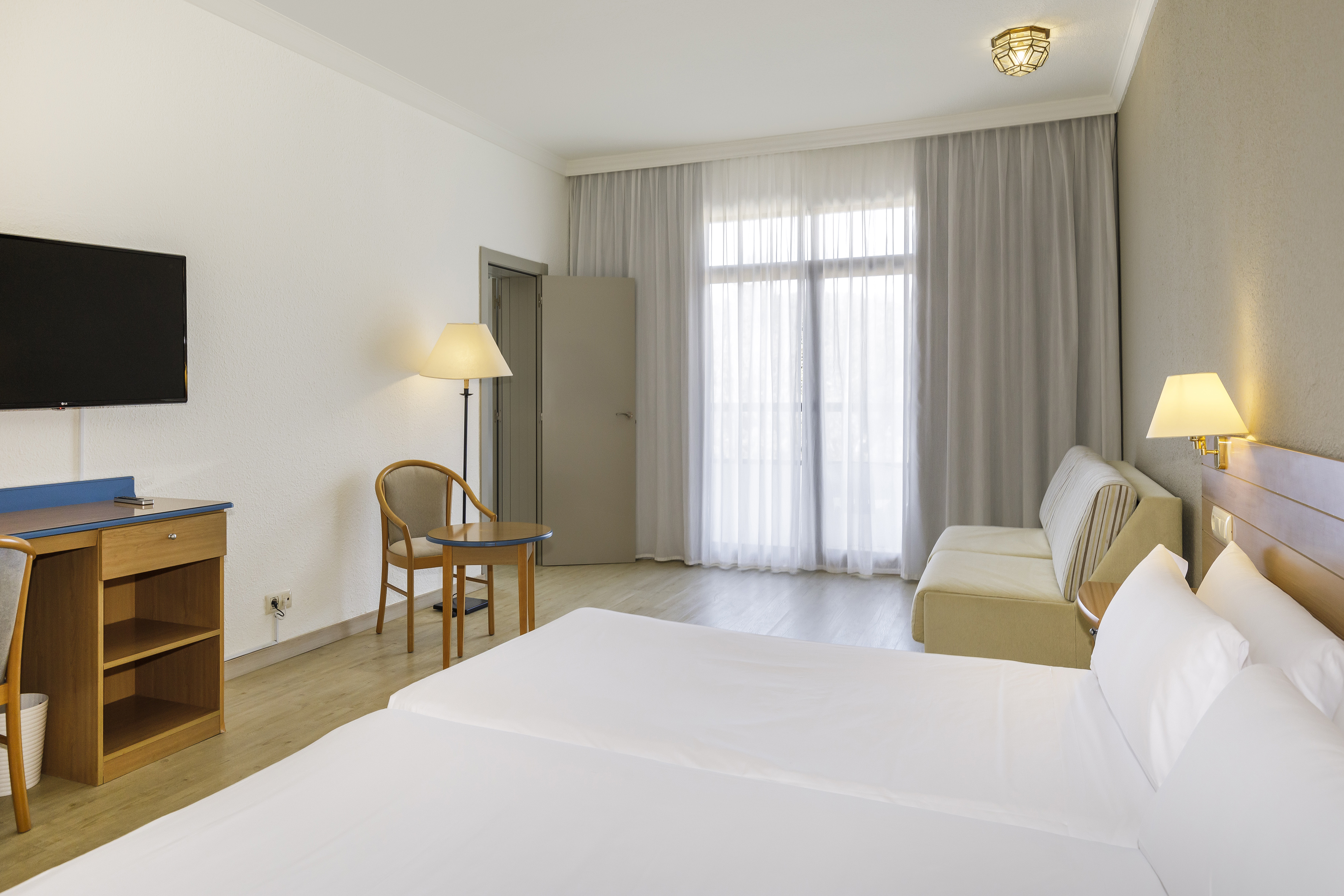 Bright hotel room with two beds, sofa, desk, and large window.