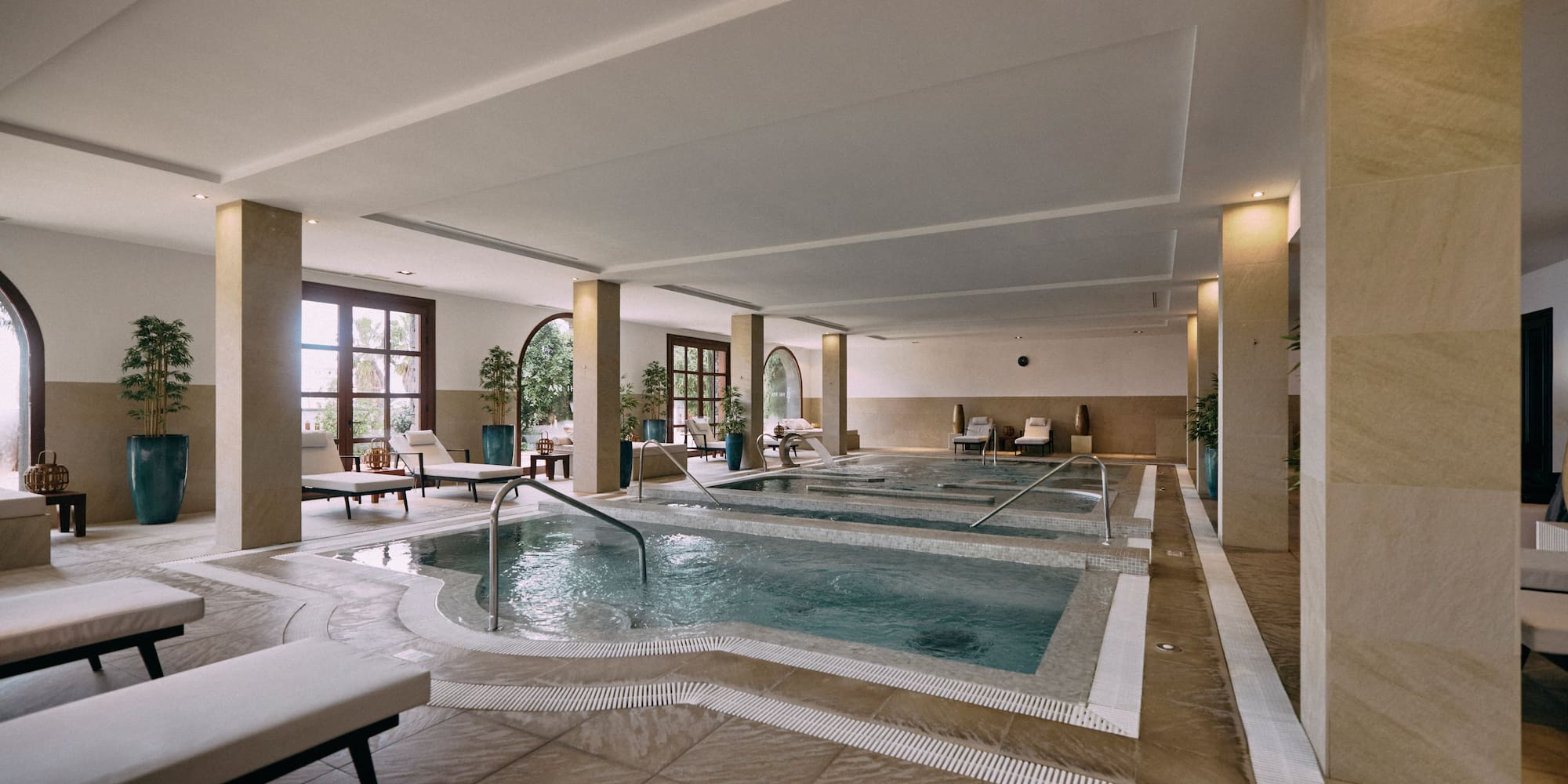 Indoor spa with multiple relaxation pools, loungers, and large arched windows.