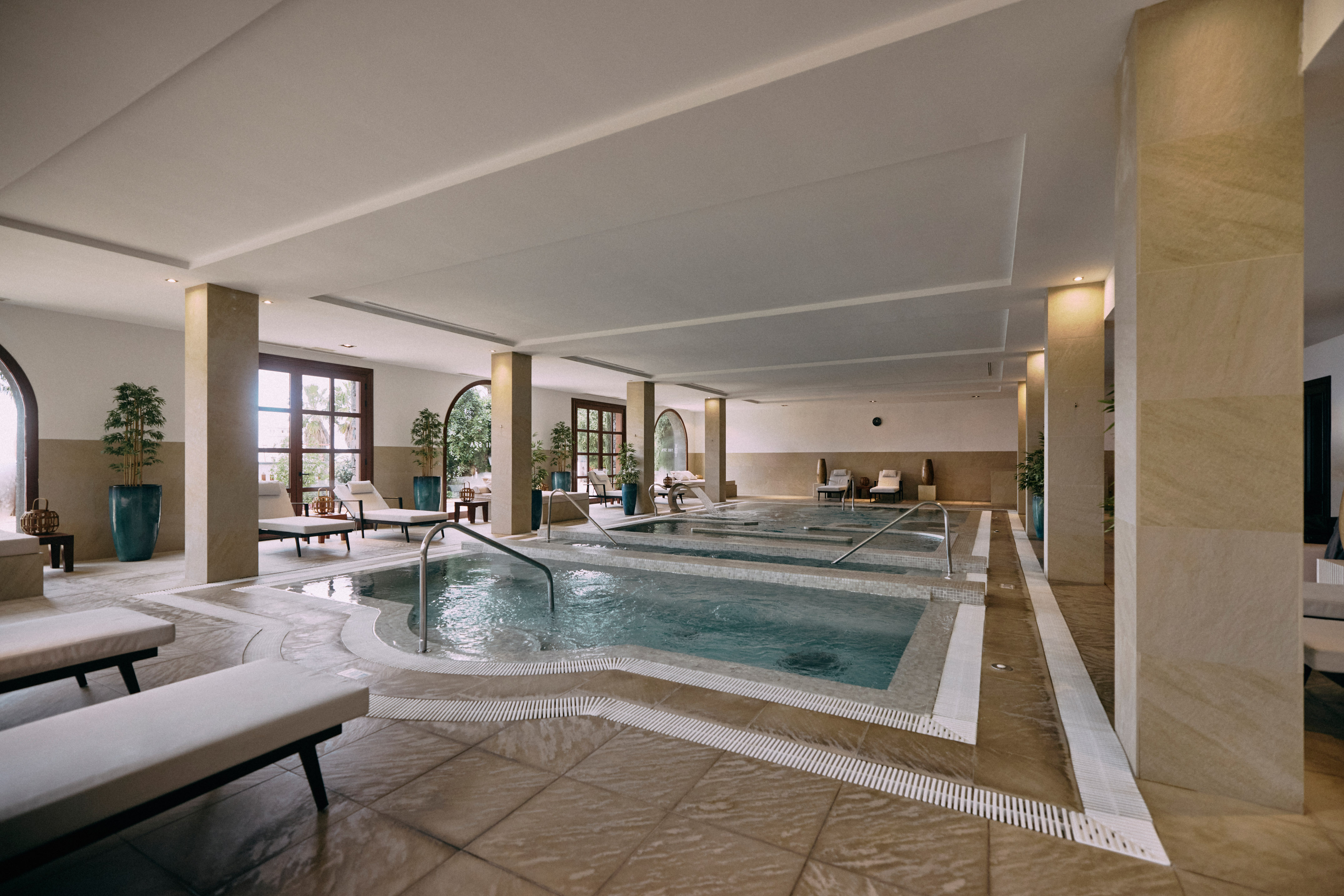 Indoor spa with multiple relaxation pools, loungers, and large arched windows.