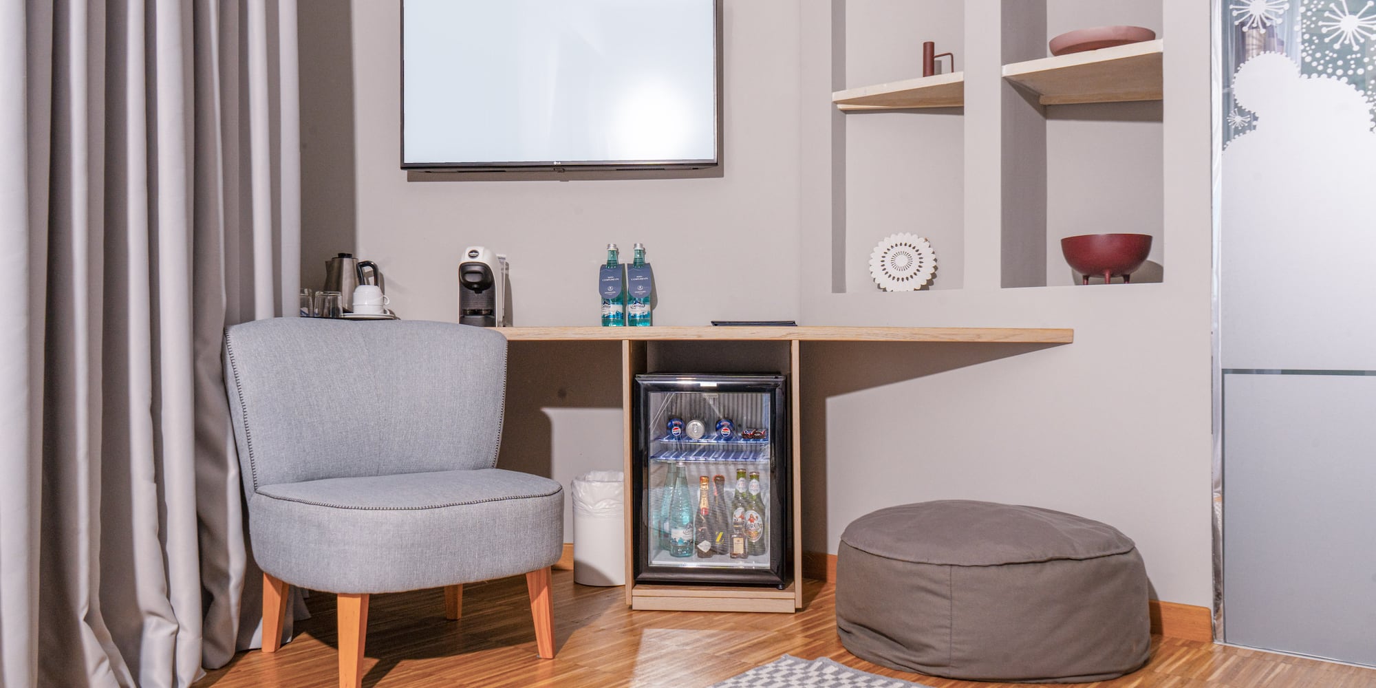 Hotel guest room with minibar, TV, grey armchair, and coffee machine.