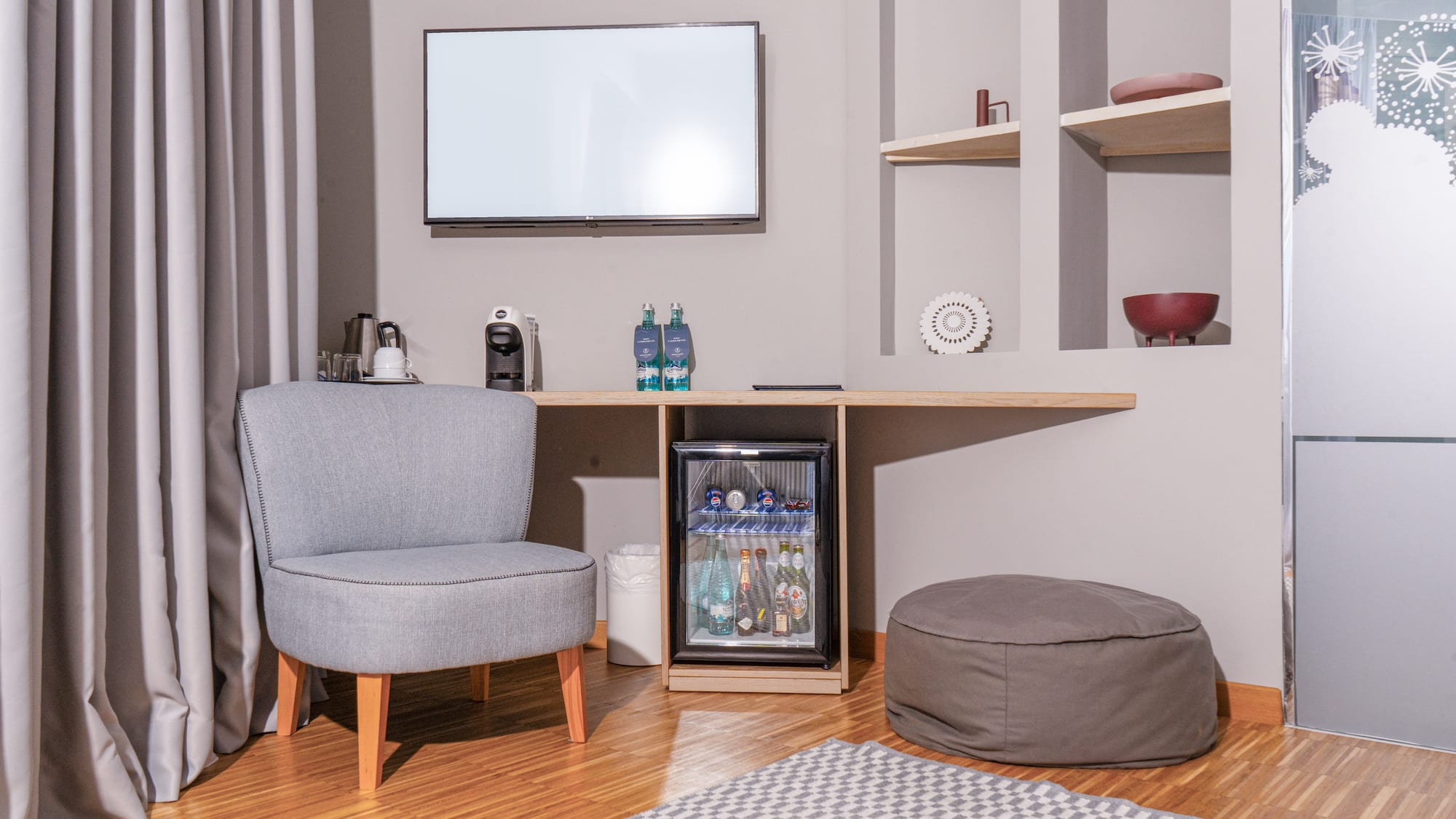 Hotel guest room with minibar, TV, grey armchair, and coffee machine.