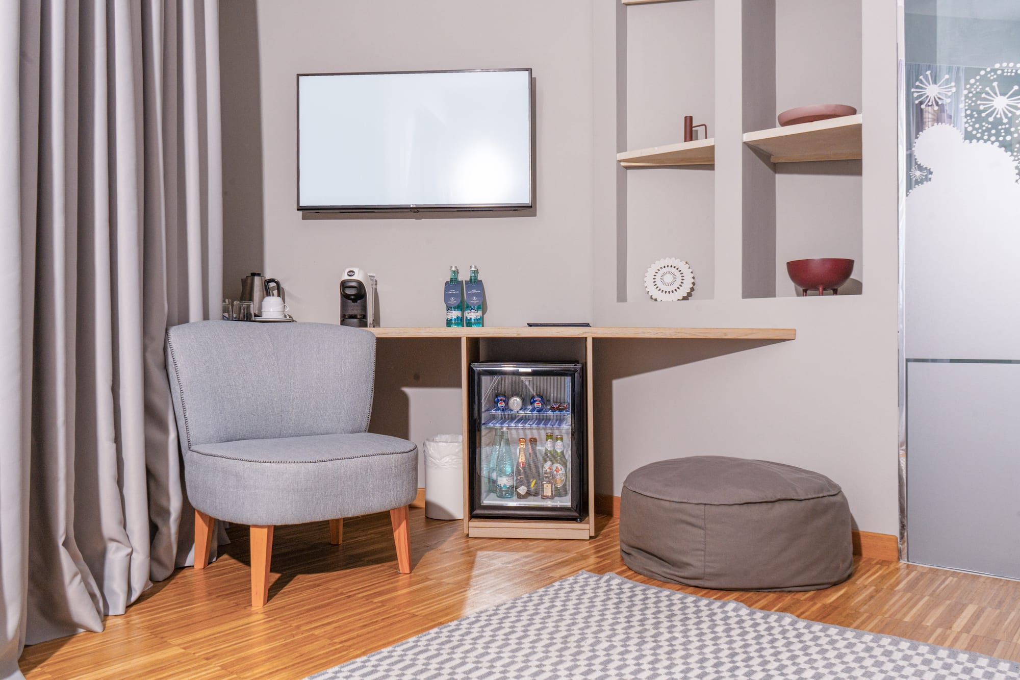 Hotel guest room with minibar, TV, grey armchair, and coffee machine.