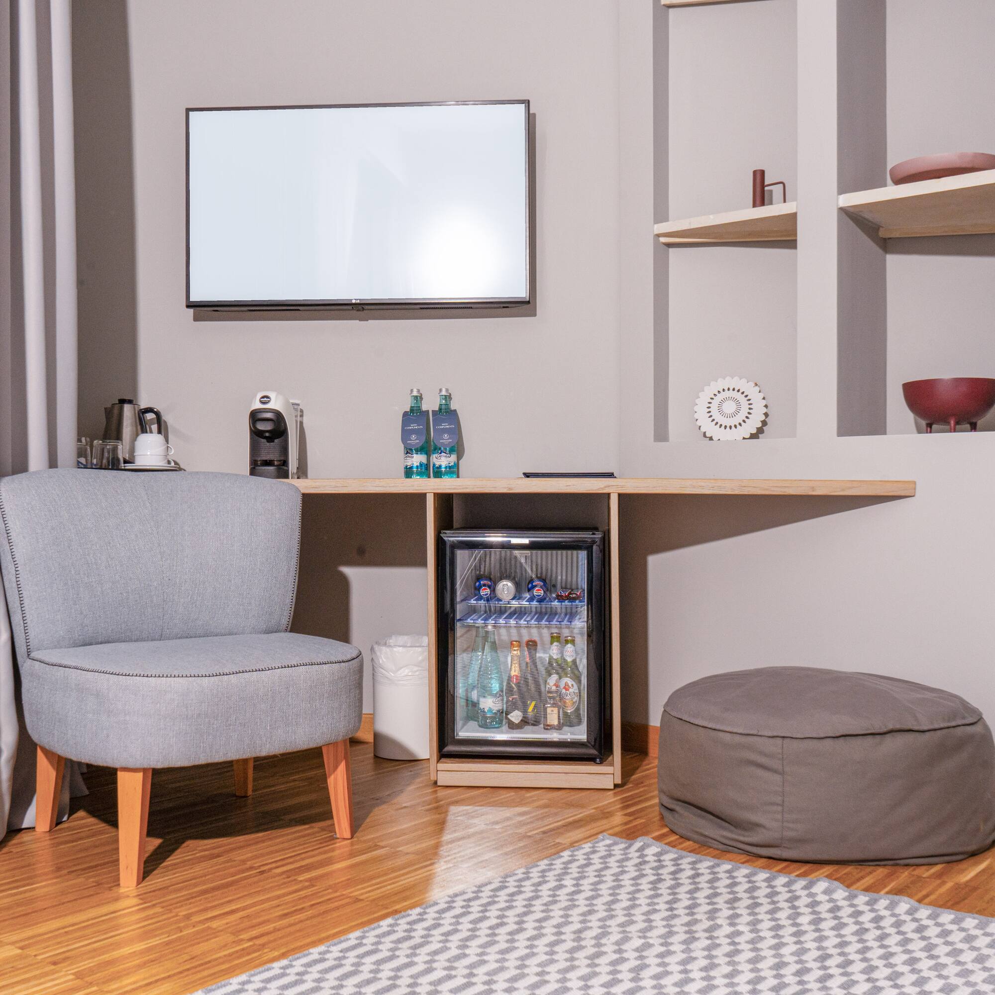 Hotel guest room with minibar, TV, grey armchair, and coffee machine.