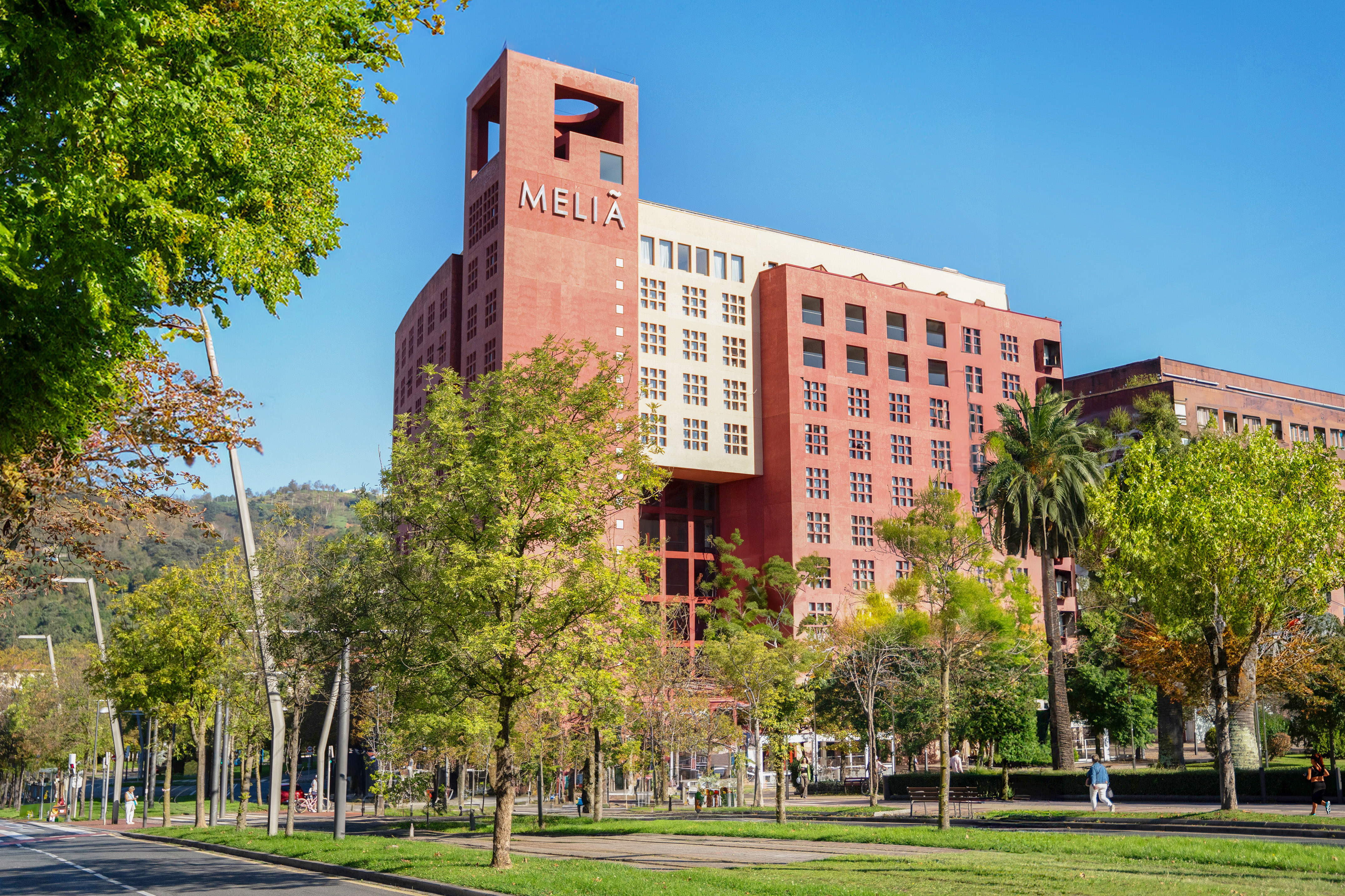 Meliá hotel building with red and white facade, green trees, and urban street.