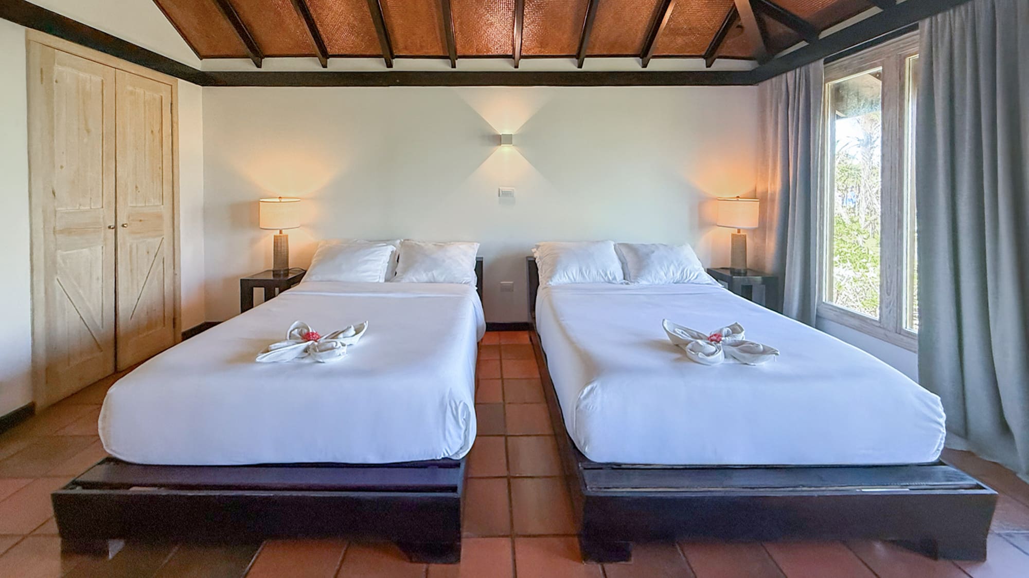 Tropical bedroom with two white beds, woven ceiling, and terracotta tile floor.