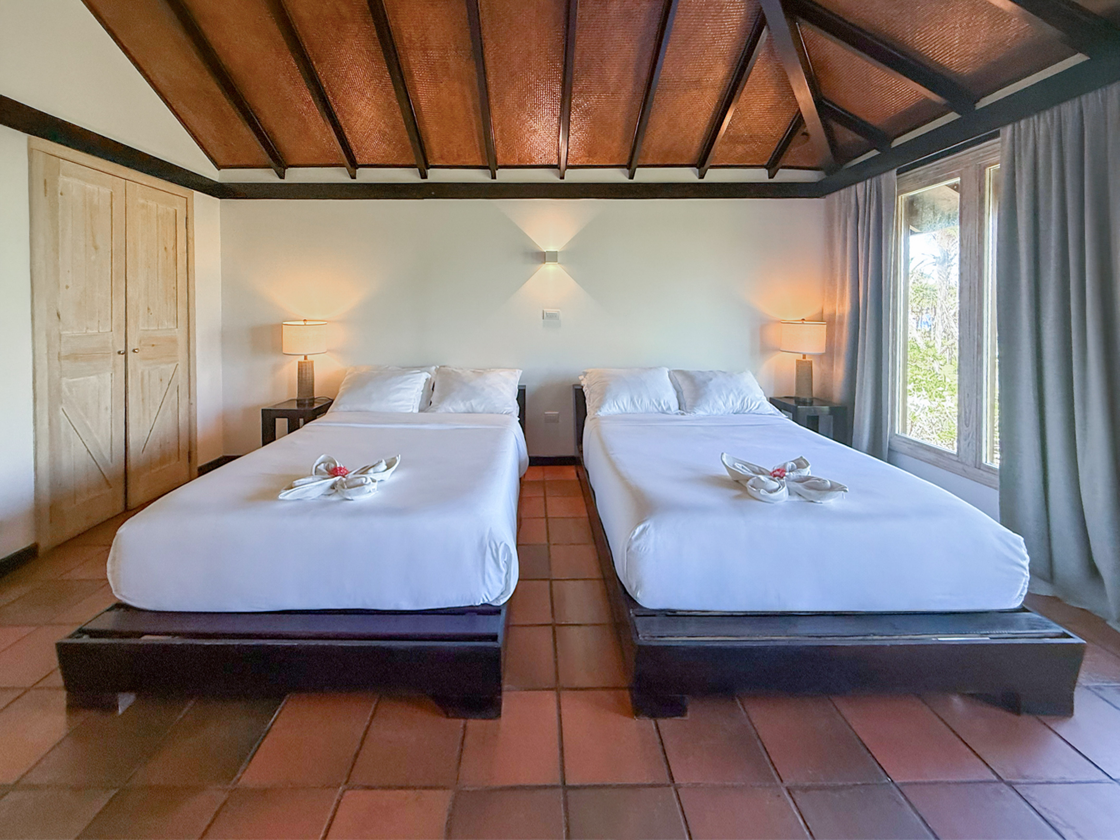 Tropical bedroom with two white beds, woven ceiling, and terracotta tile floor.