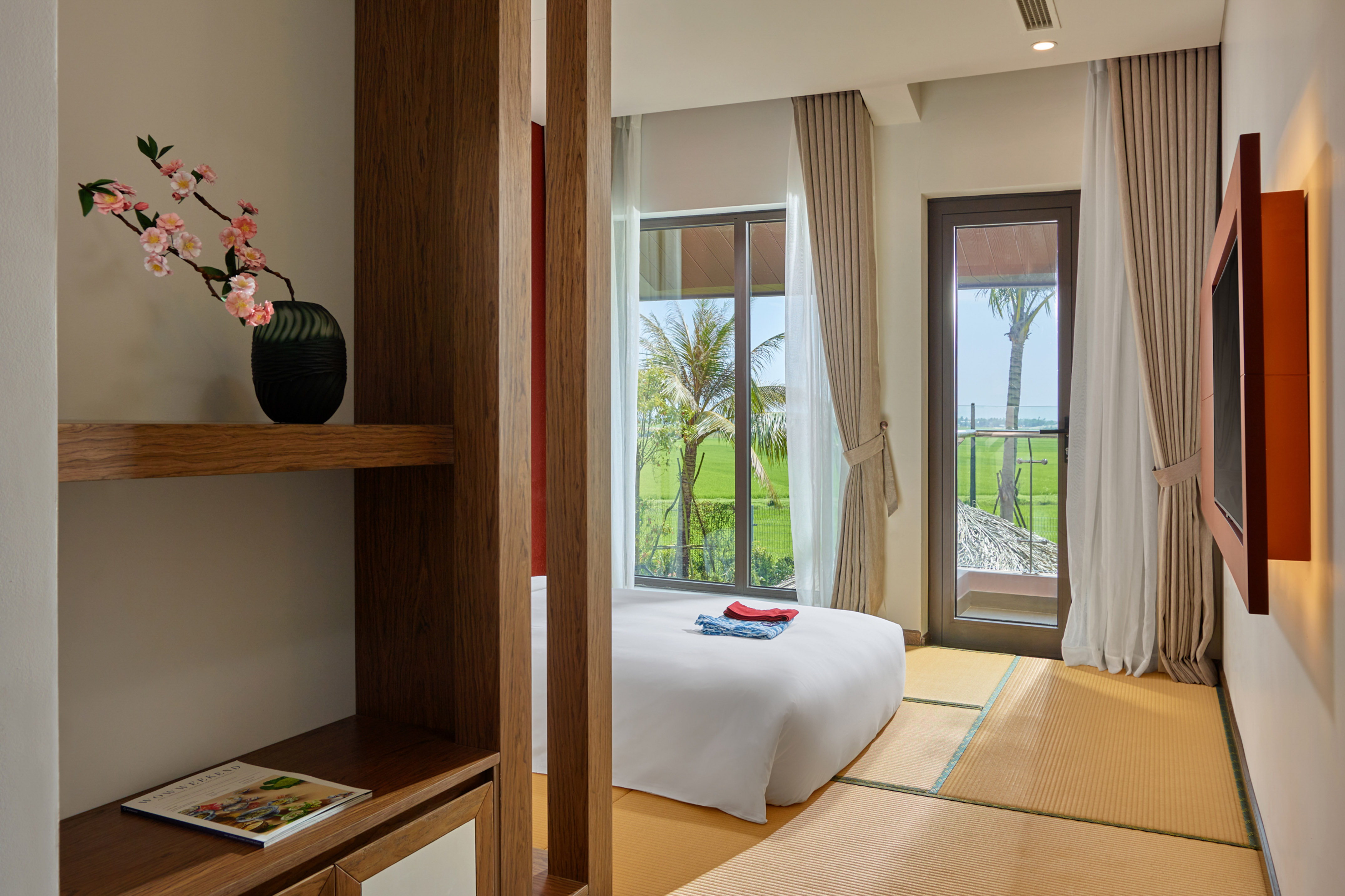 Modern bedroom with tatami floor, white bed, and rice field view with palm trees.